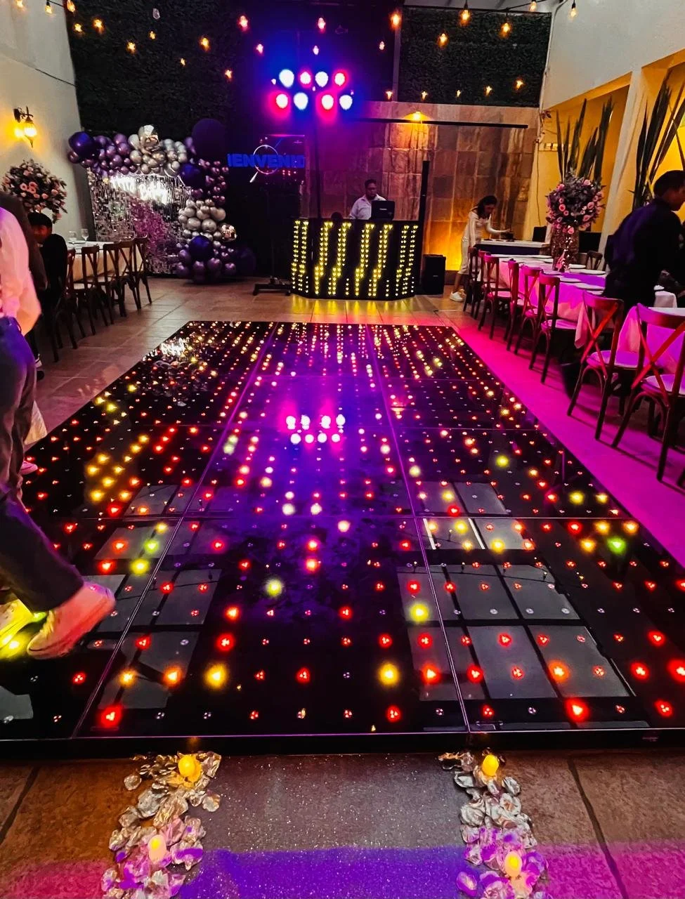 Pista de baile iluminada con luces de colores en celebración, DJ en plataforma con decoraciones de globos y flores, mesas con arreglos florales y ambiente festivo.