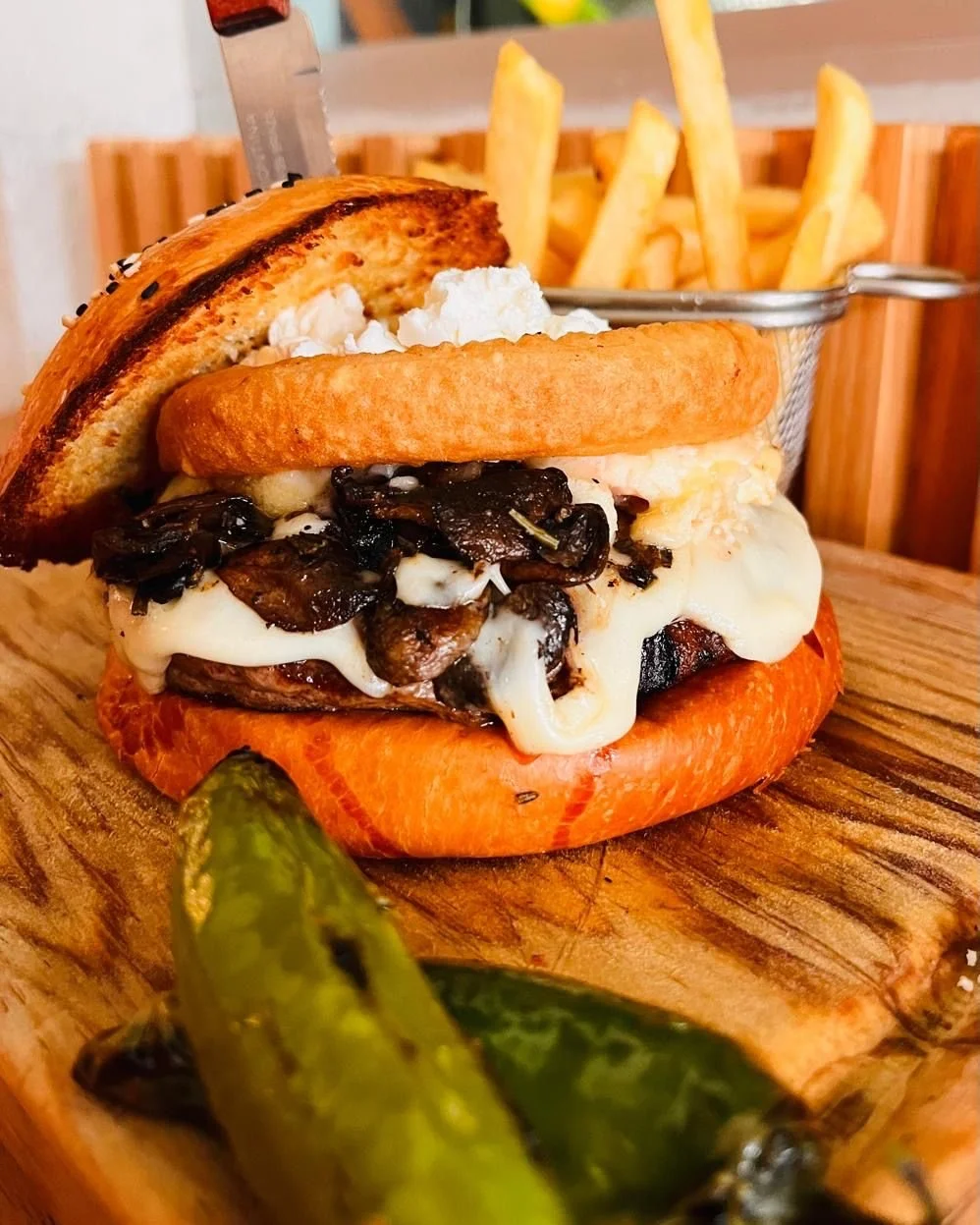 Hamburguesa con queso, champiñones, tocino, pan tostado, papas fritas en un recipiente metálico, y un chile jalapeño en primer plano.
