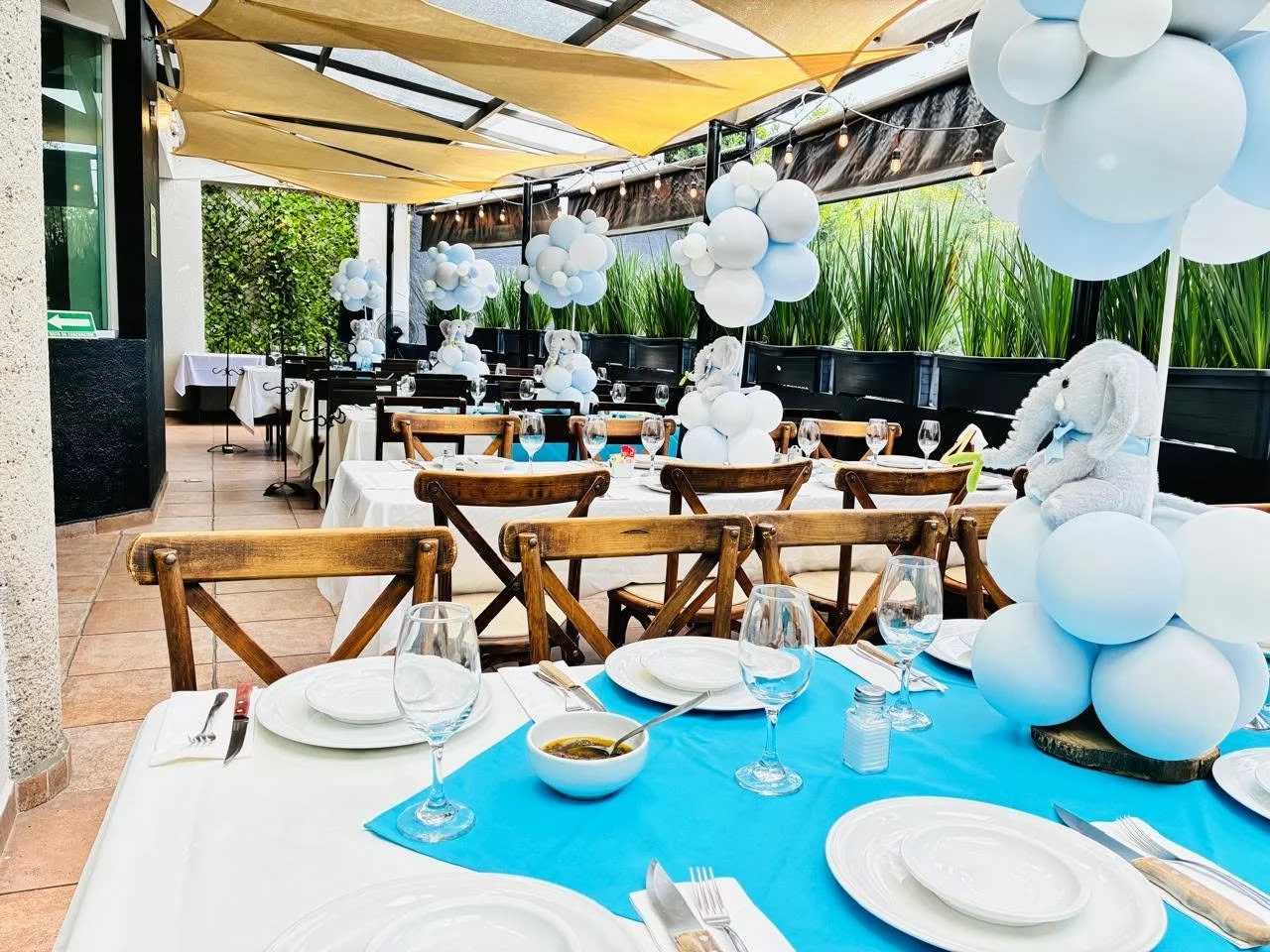 Decoración para evento infantil con bolas blancas, ositos de peluche y un tema de elefantes en una terraza cubierta con toldos beige y luces decorativas.