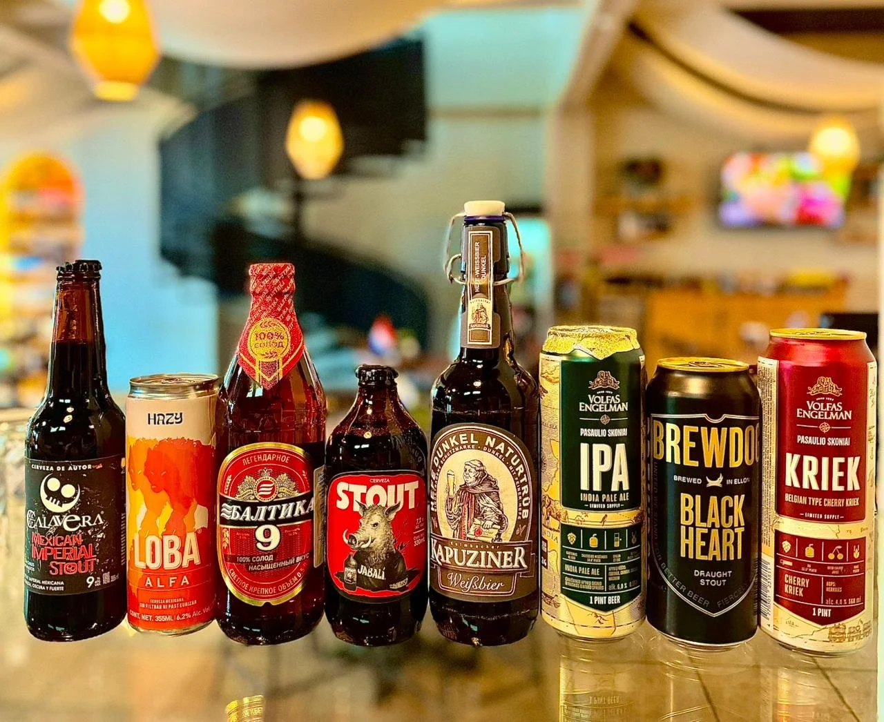 Variedad de latas y botellas de cerveza distintas en una mesa, con fondo de interior de un bar o restaurante.