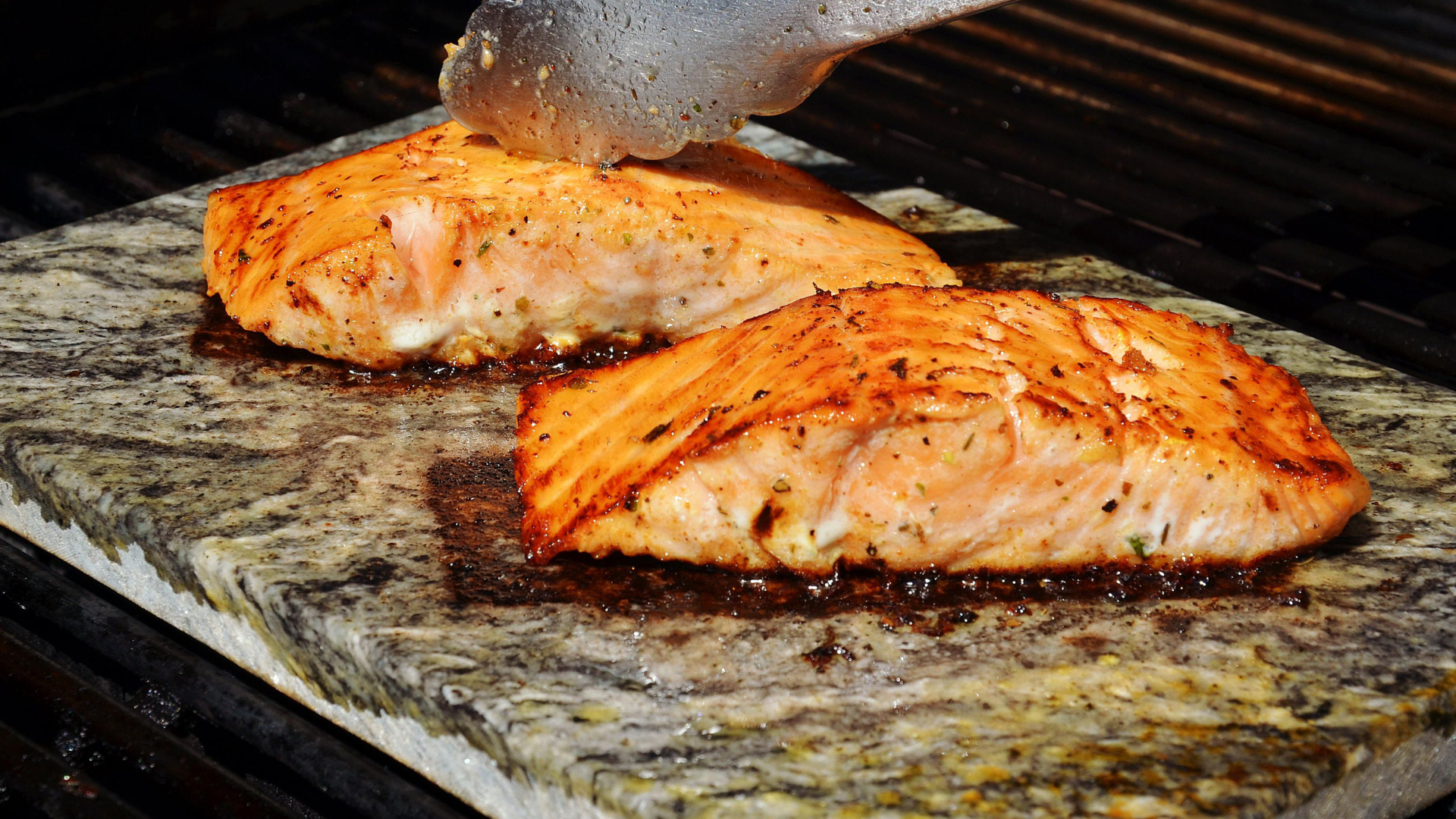 Salmón a la parrilla en una superficie de piedra, siendo cocinado con una espátula, con marcas de grill y aceite.