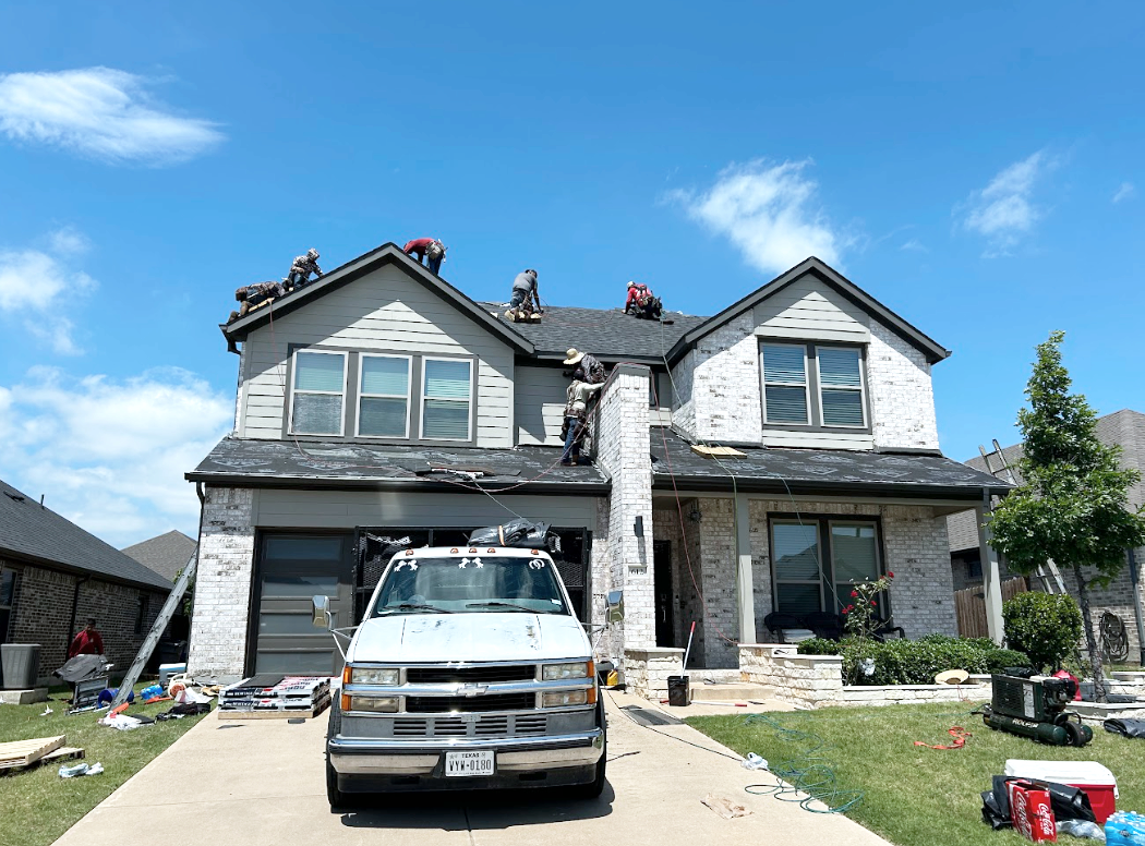 Construction workers repairing a roof on a two-story house, with tools and equipment on the driveway and lawn, and a blue sky with some clouds.