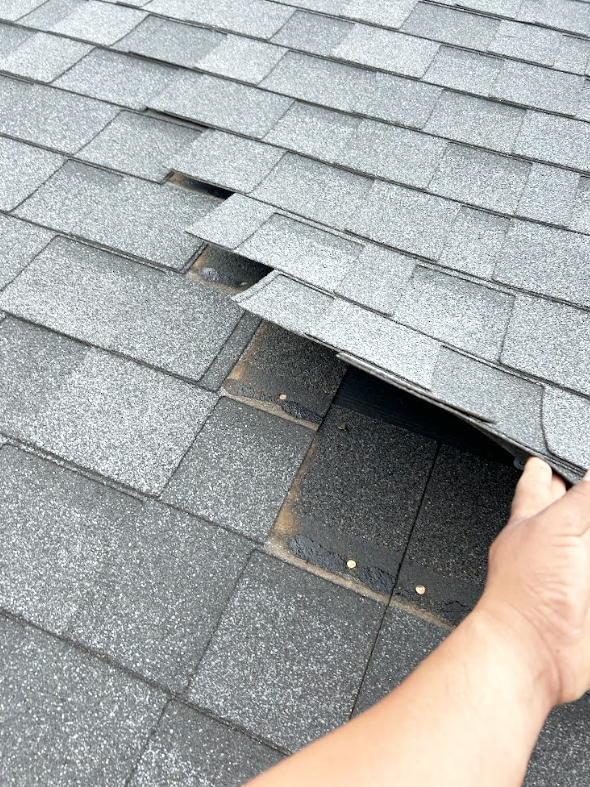 Person inspecting damaged asphalt shingles on a roof, revealing a gap or missing shingles.