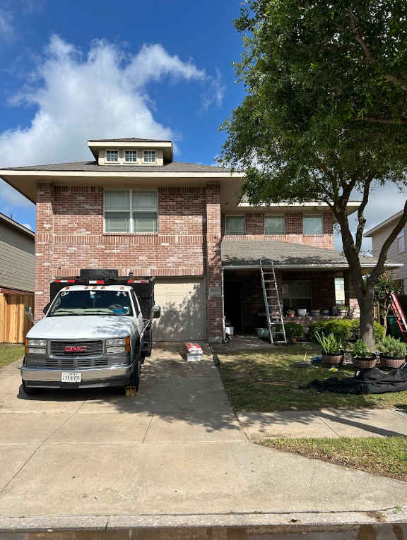 Two-story brick house with a vehicle parked in the driveway, a ladder leaning against the house, and potted plants near the porch. There's a tree on the right side, and the sky is partly cloudy.