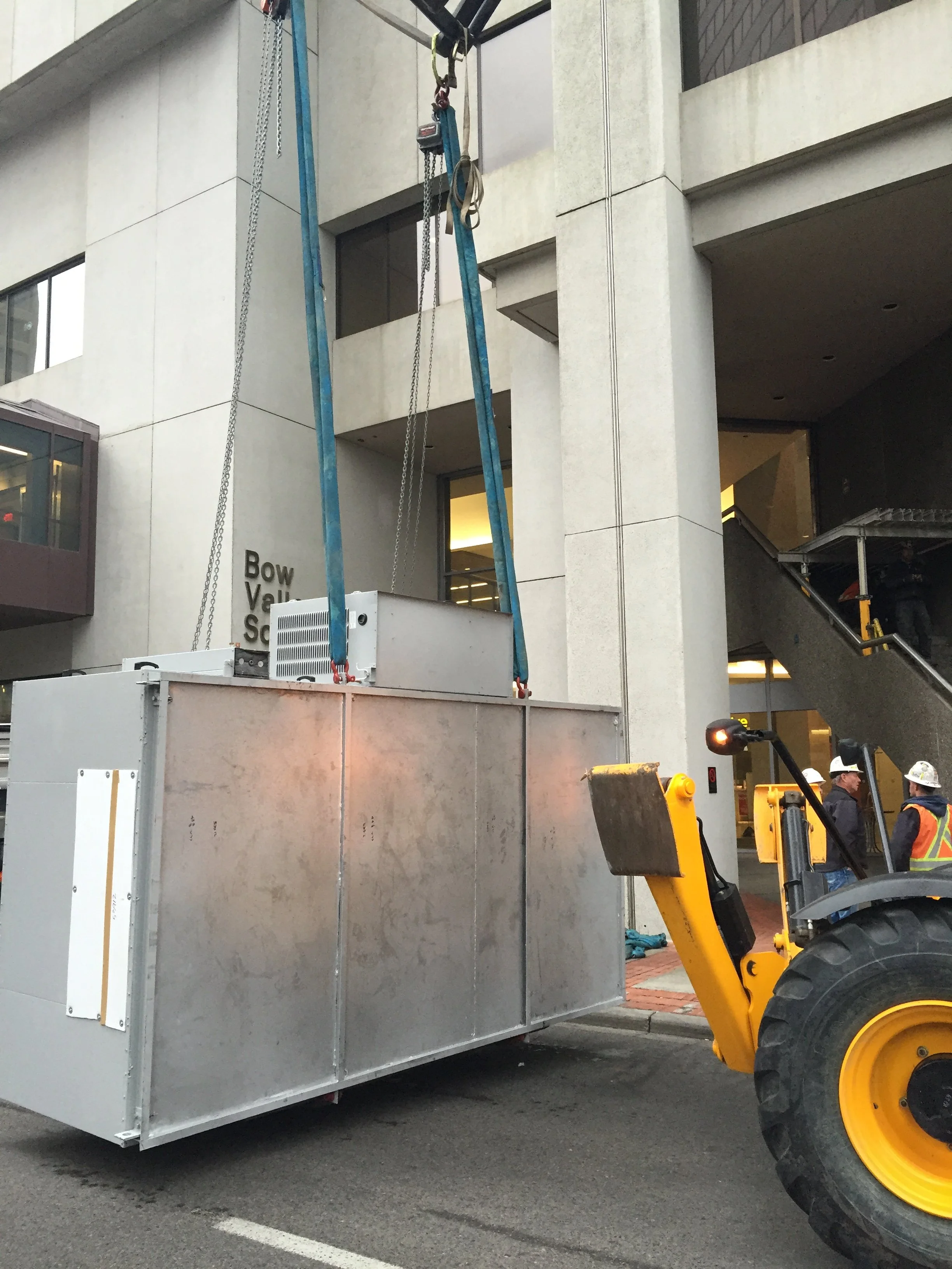Construction workers operating heavy machinery lifting a large, metallic modular container in an urban area.