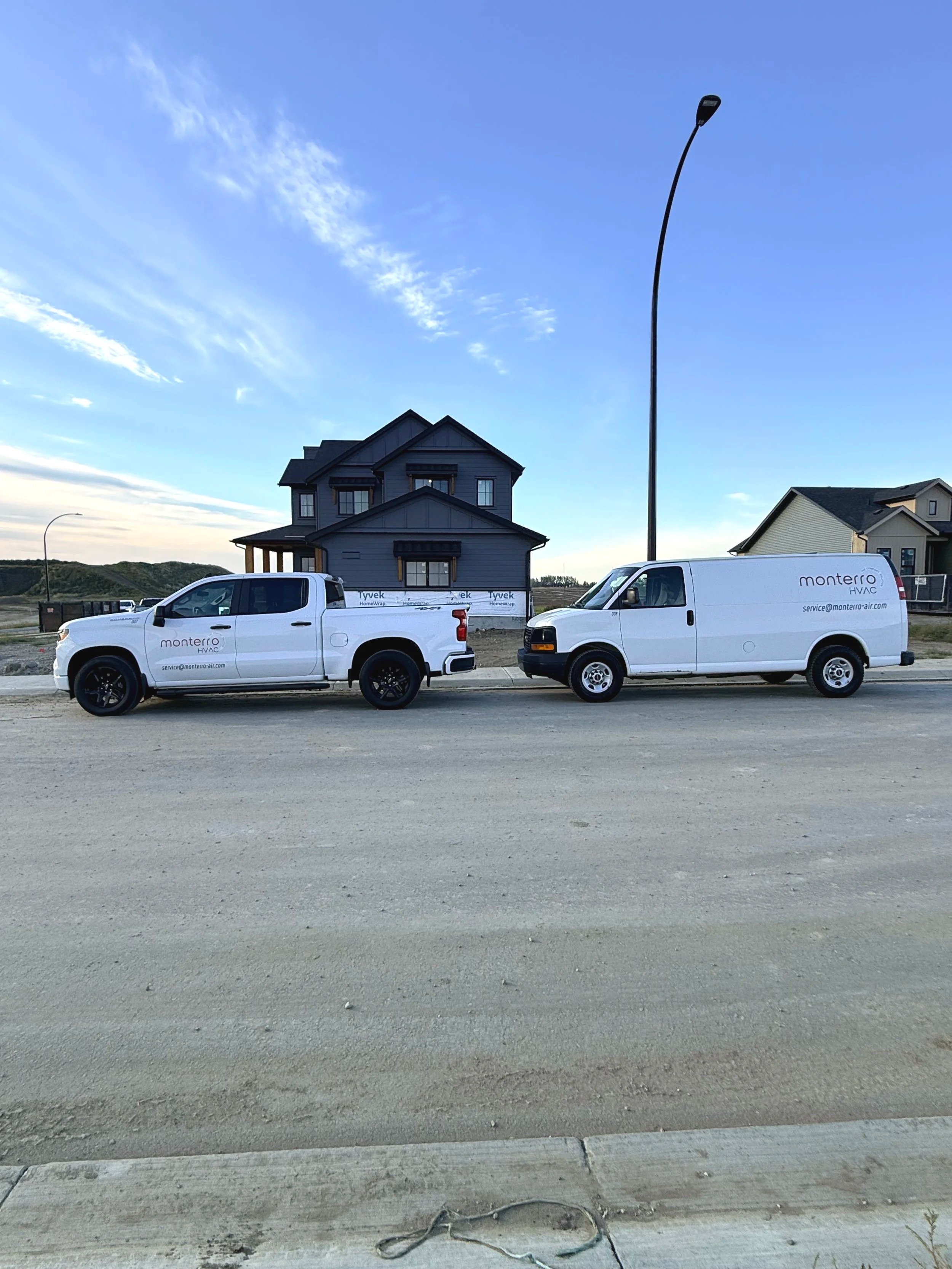 Two white service trucks parked on a street in front of a blue house, with a clear sky and a streetlamp in the background.