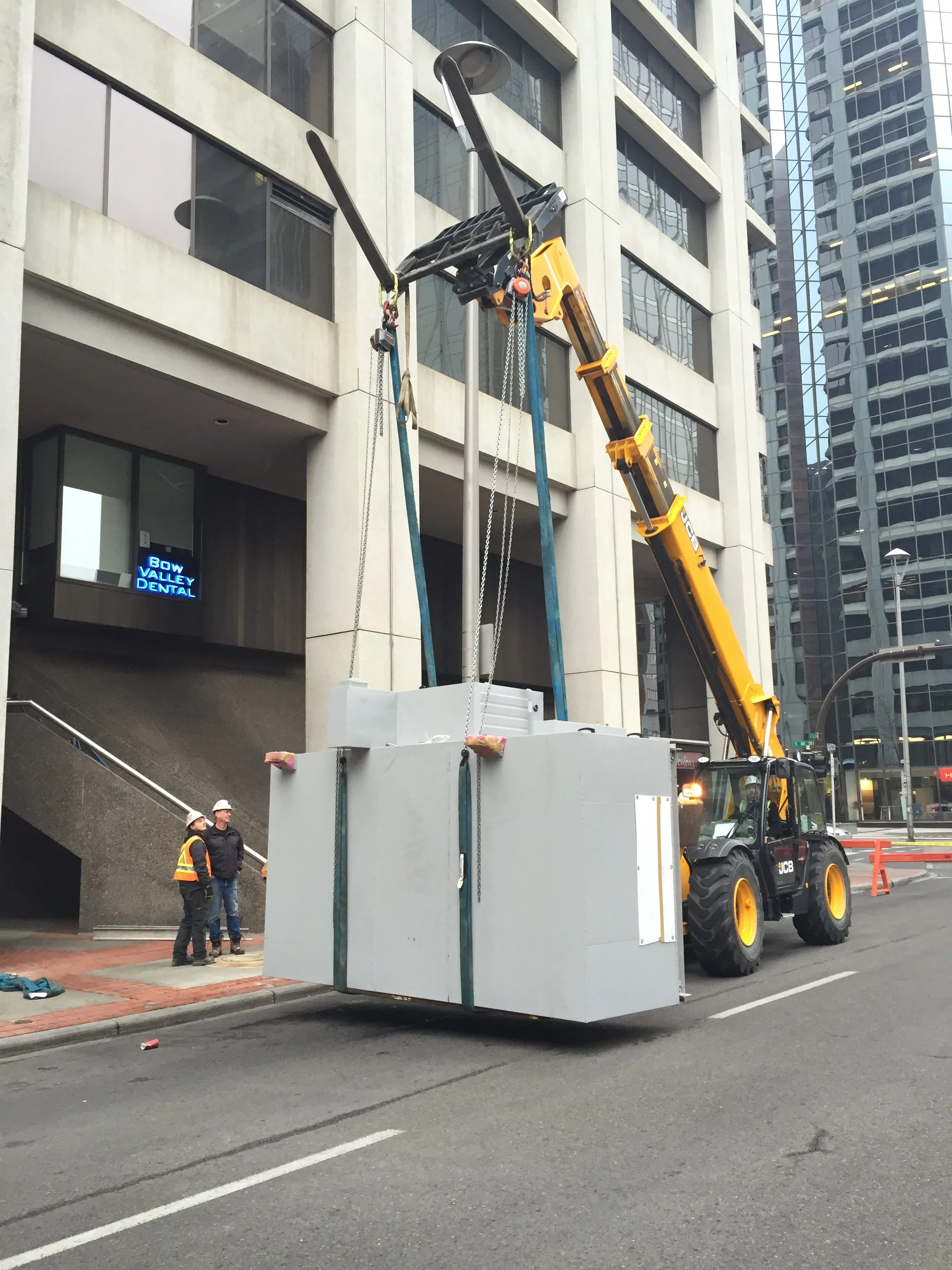 Construction workers in safety vests and helmets stand near a large crane lifting a white modular building or container on a city street. Tall office buildings are in the background, and there is a dental office named Bow Valley Dental visible on a lower floor.