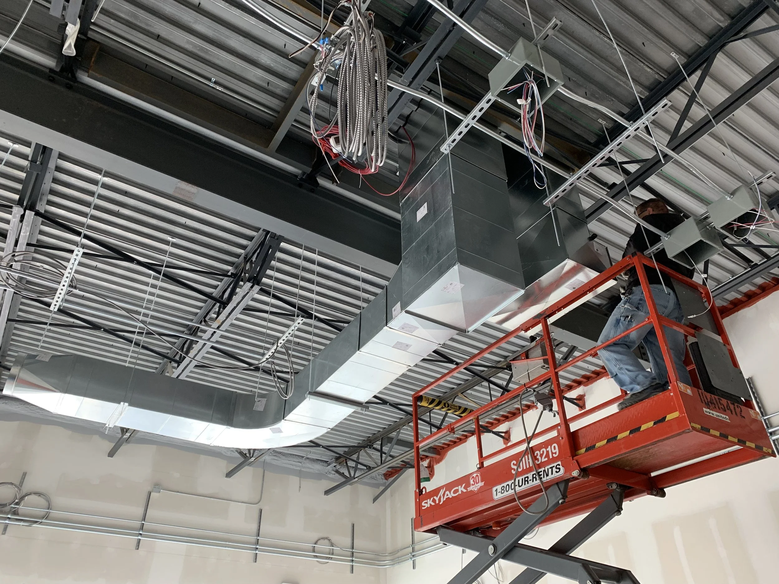 Construction worker on red scissor lift installing ceiling ducts and wiring in an unfinished building ceiling.