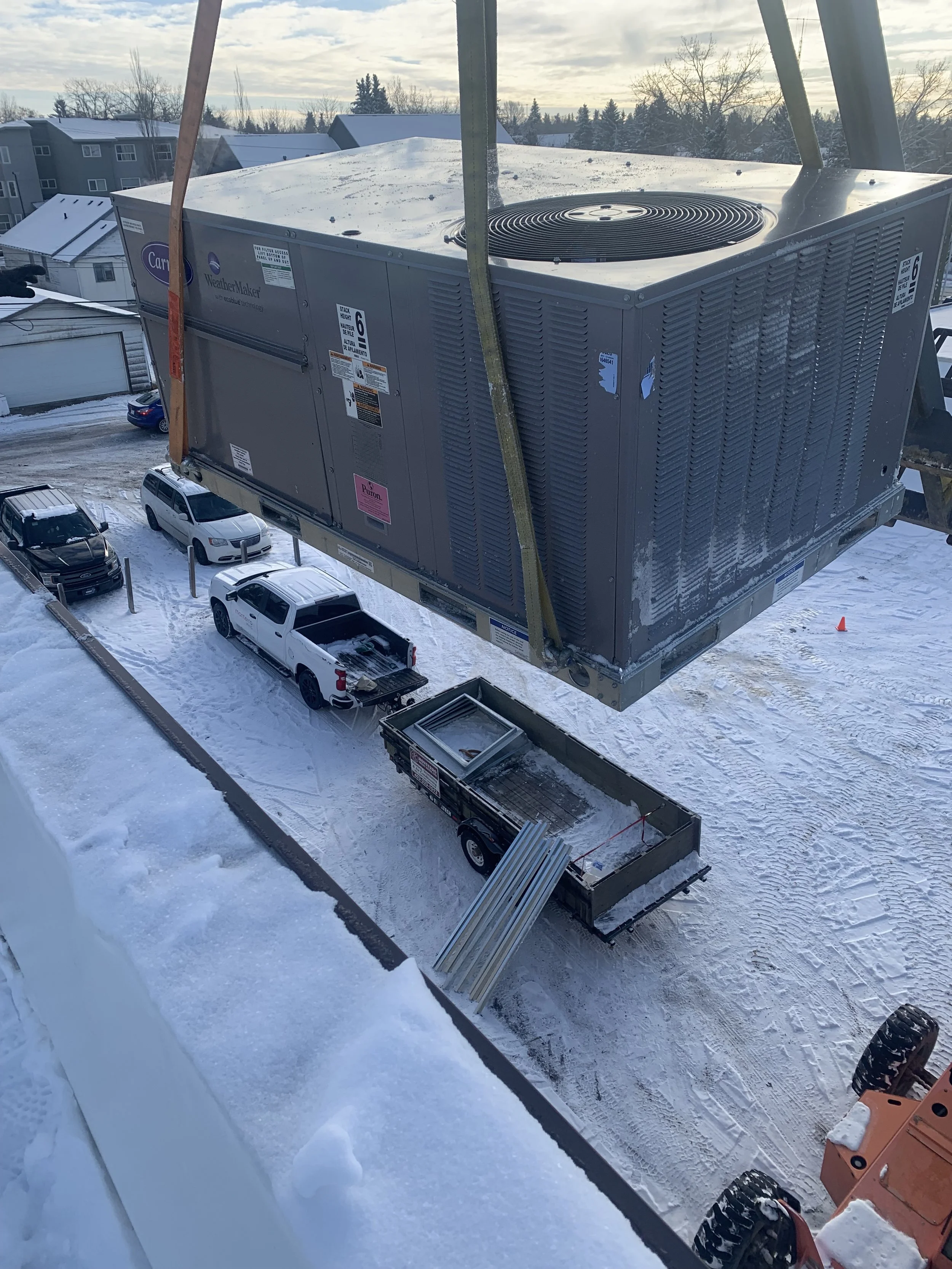A rooftop HVAC unit installed on a building in a snowy residential area with parked cars below, overcast sky, and leafless trees in the background.