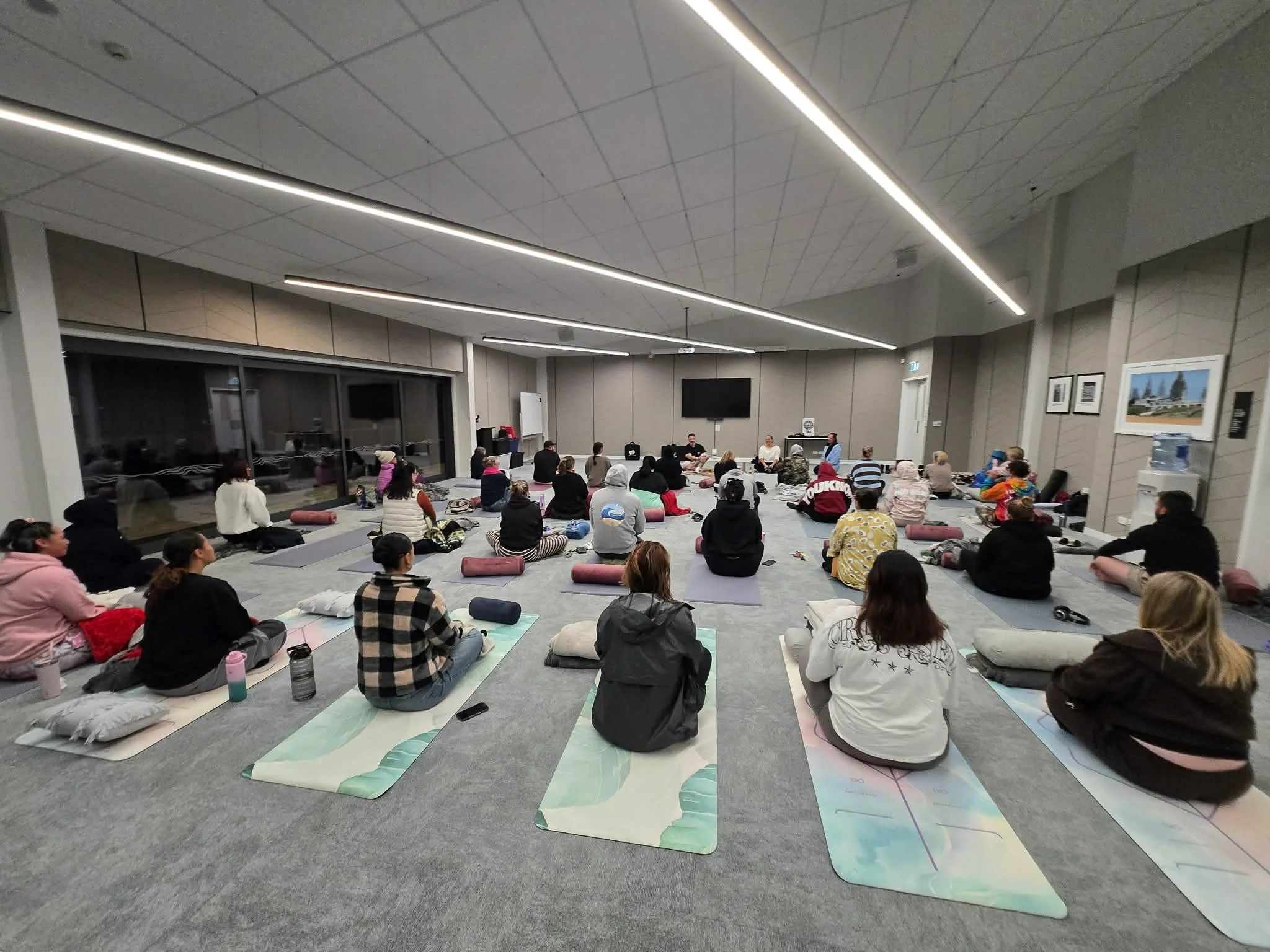A large group of people sitting on yoga mats in a spacious room, attending a meditation or yoga class, with some looking at the front where an instructor may be leading the session.