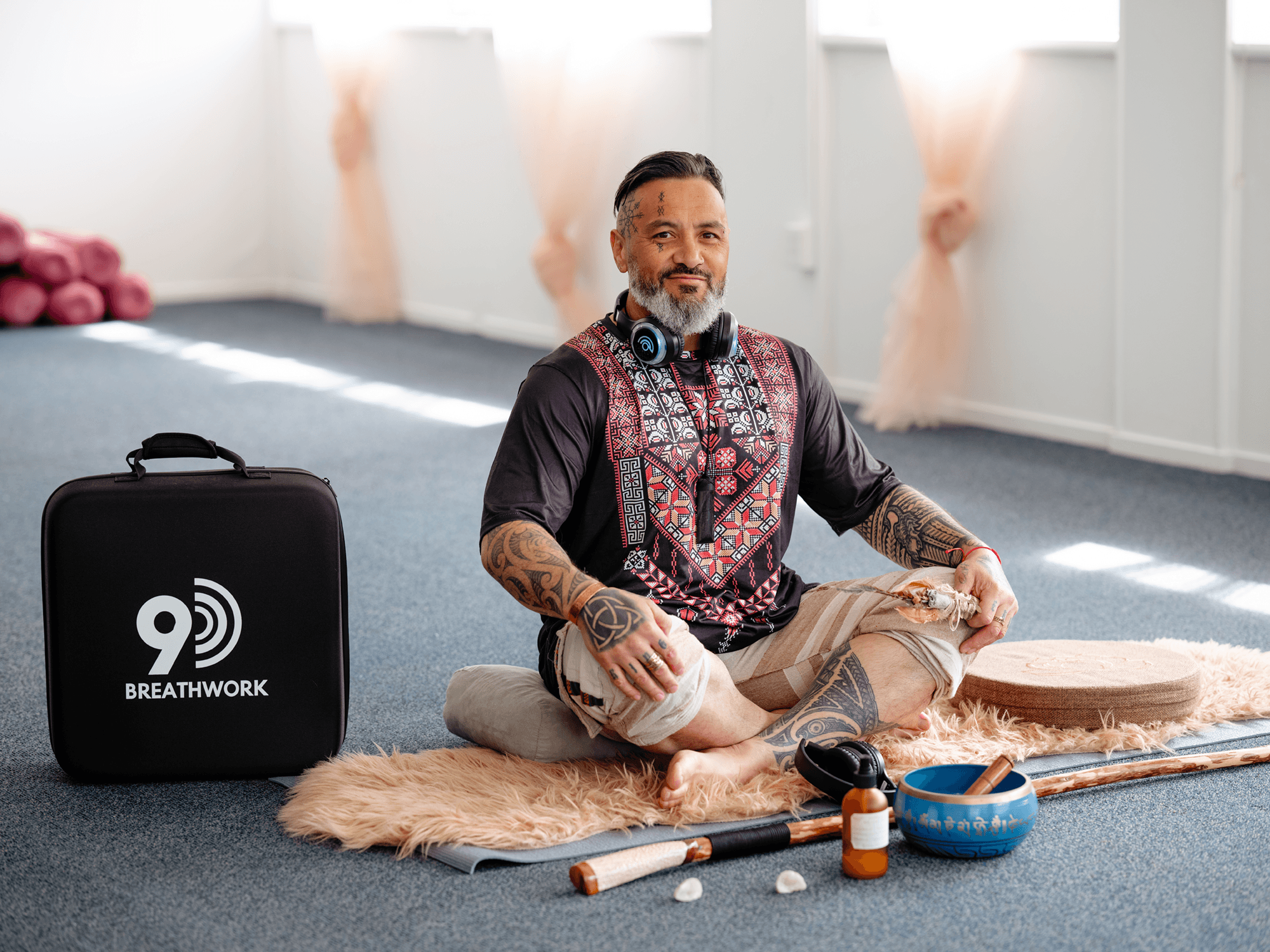 A man with tattoos around his face and arms, gray beard, wearing a traditional designed shirt, sitting cross-legged on a furry rug at an indoor yoga studio. Yoga props, a large black speaker with '9 BREATHWORK' written on it, and a bowl with various objects are nearby. The background shows a bright, clean room with natural light coming through windows and pink yoga mats rolled up against the wall.
