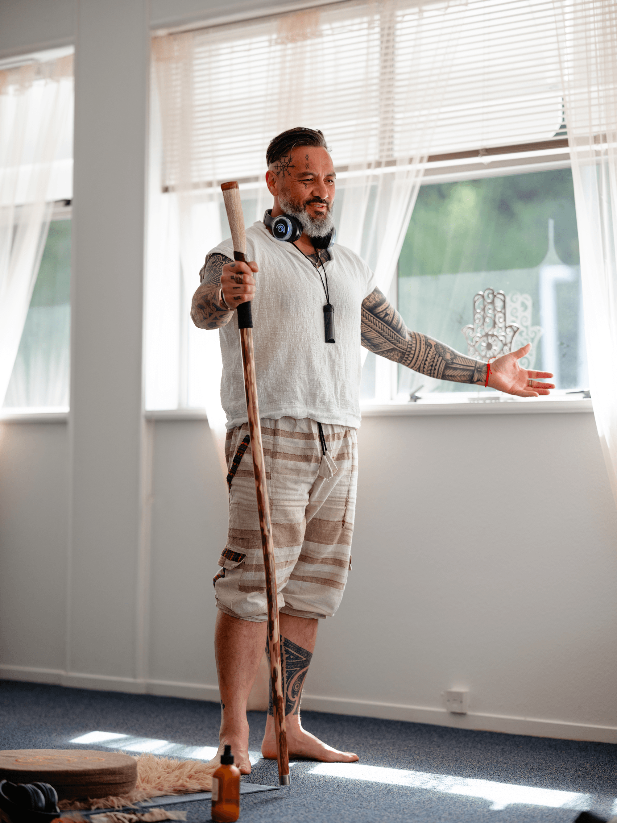 A man with tattoos, a gray beard, and a tattooed prayer symbol on his head is standing barefoot in a brightly lit room, holding a walking stick and gesturing with his other hand. He is wearing a white shirt, beige shorts, and has headphones around his neck. The room has large windows with blinds and sheer curtains, with some yoga mats, a bottle of lotion, and a blanket on the floor.