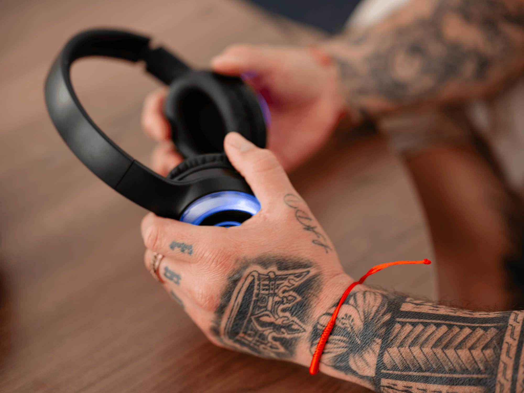 Close-up of a tattooed arm holding a black wireless gaming headset with blue lighting, resting on a wooden surface with an orange cord nearby.