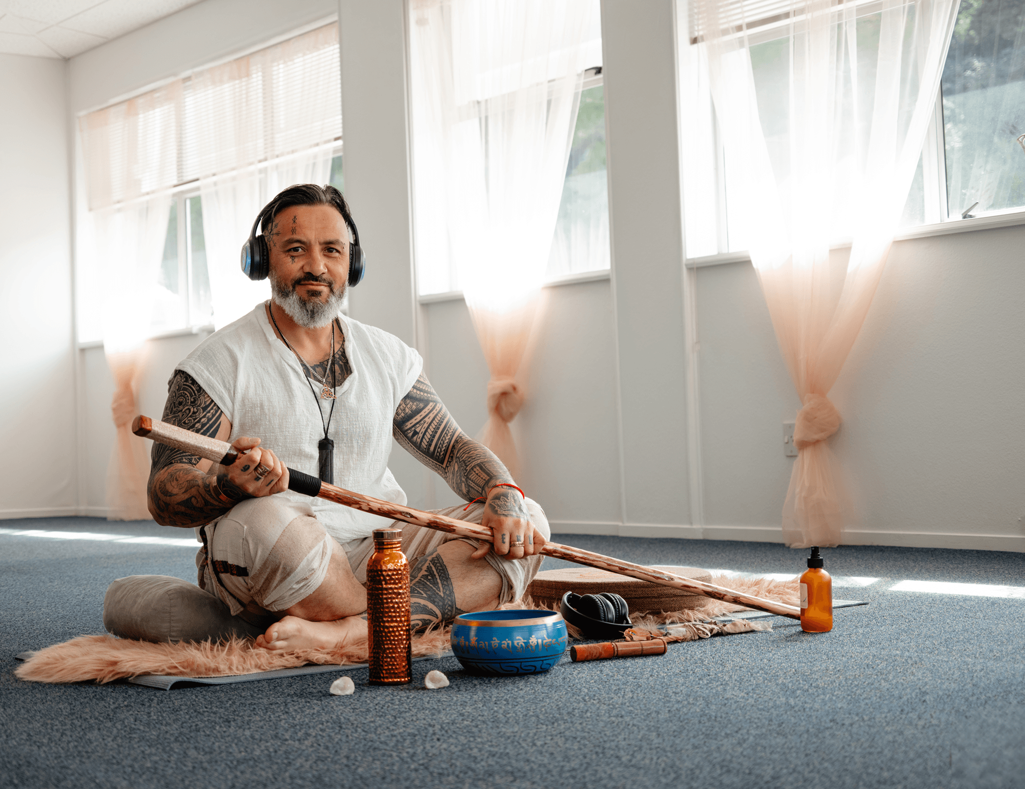 Man with tattoos, beard, and headphones sitting on a fluffy rug with spiritual items, including bowls, bottles, and a mala, in a bright room with large windows and sheer curtains.