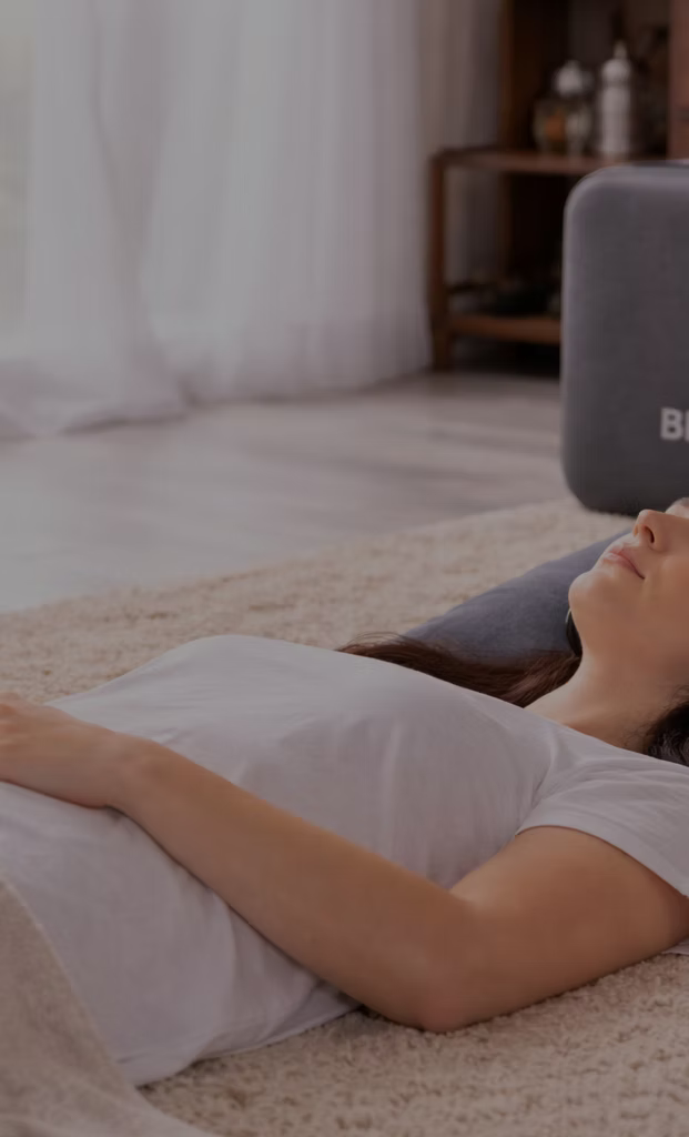 A woman lying on her back on a carpeted floor with her eyes closed, resting in a cozy living room with a gray speaker nearby and a curtain in the background.
