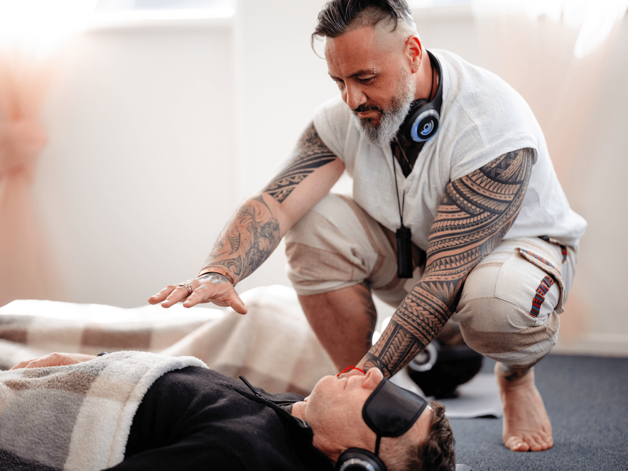 A man with tattoos and a beard, wearing headphones, kneels beside a person lying on the floor. The lying person is wearing glasses and a fleece jacket, appearing to be receiving assistance or medical help in a bright indoor setting.