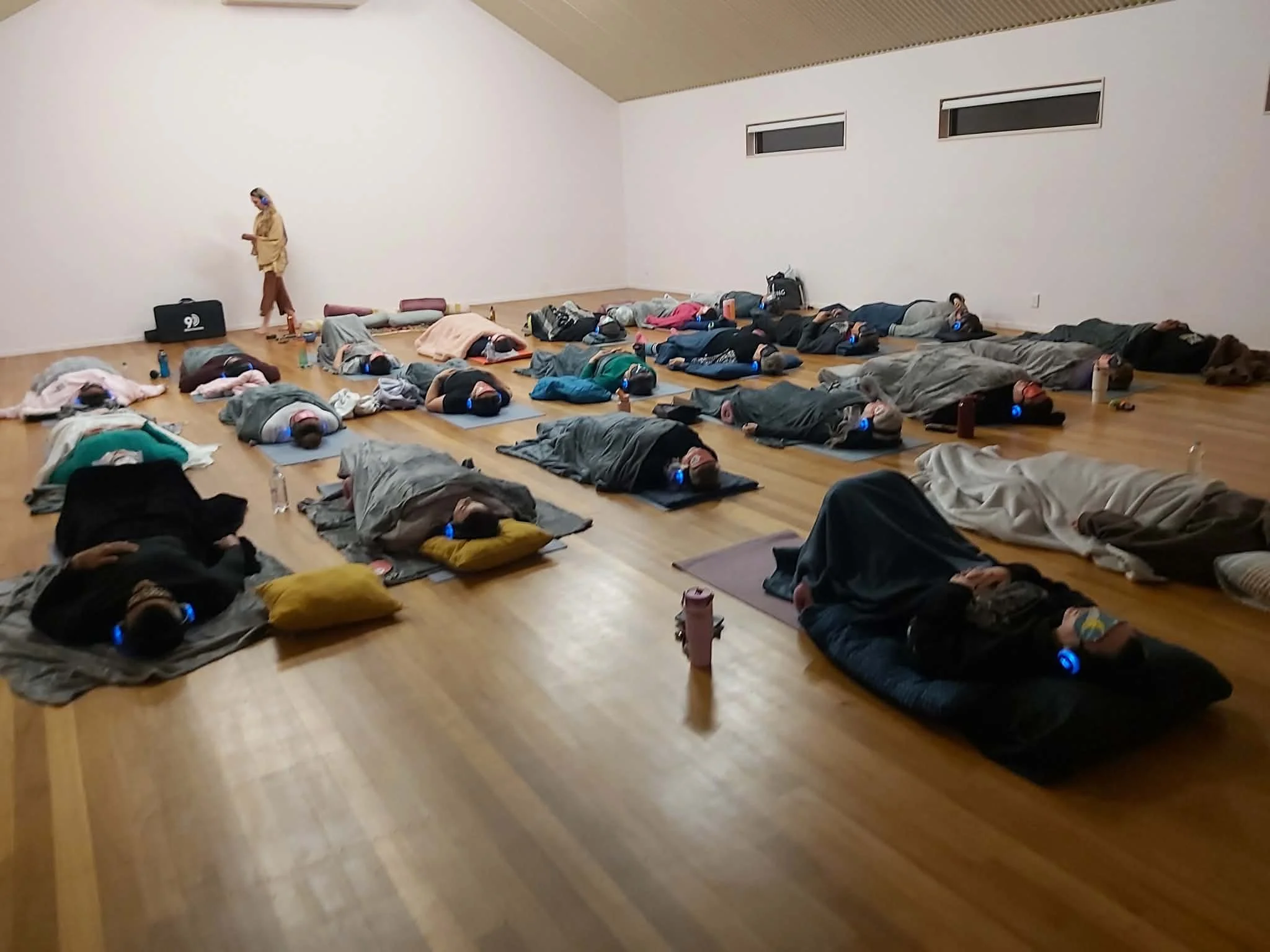 Group of people lying on yoga mats in a spacious room, participating in a meditation or yoga class, with a instructor standing at the front.