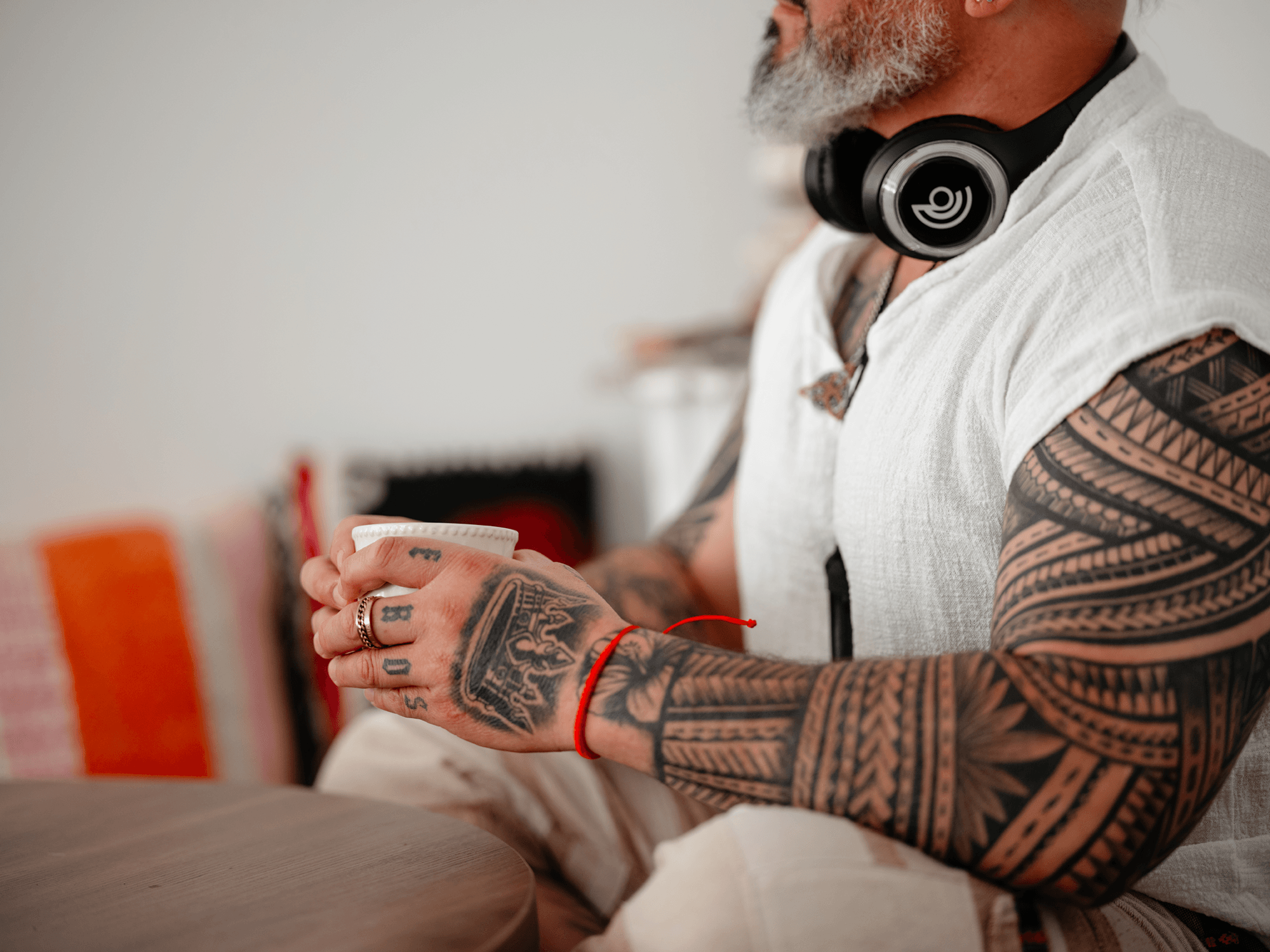 Close-up of a tattooed man wearing headphones, holding a cup with both hands, and wearing a white shirt with rolled-up sleeves, seated at a table.