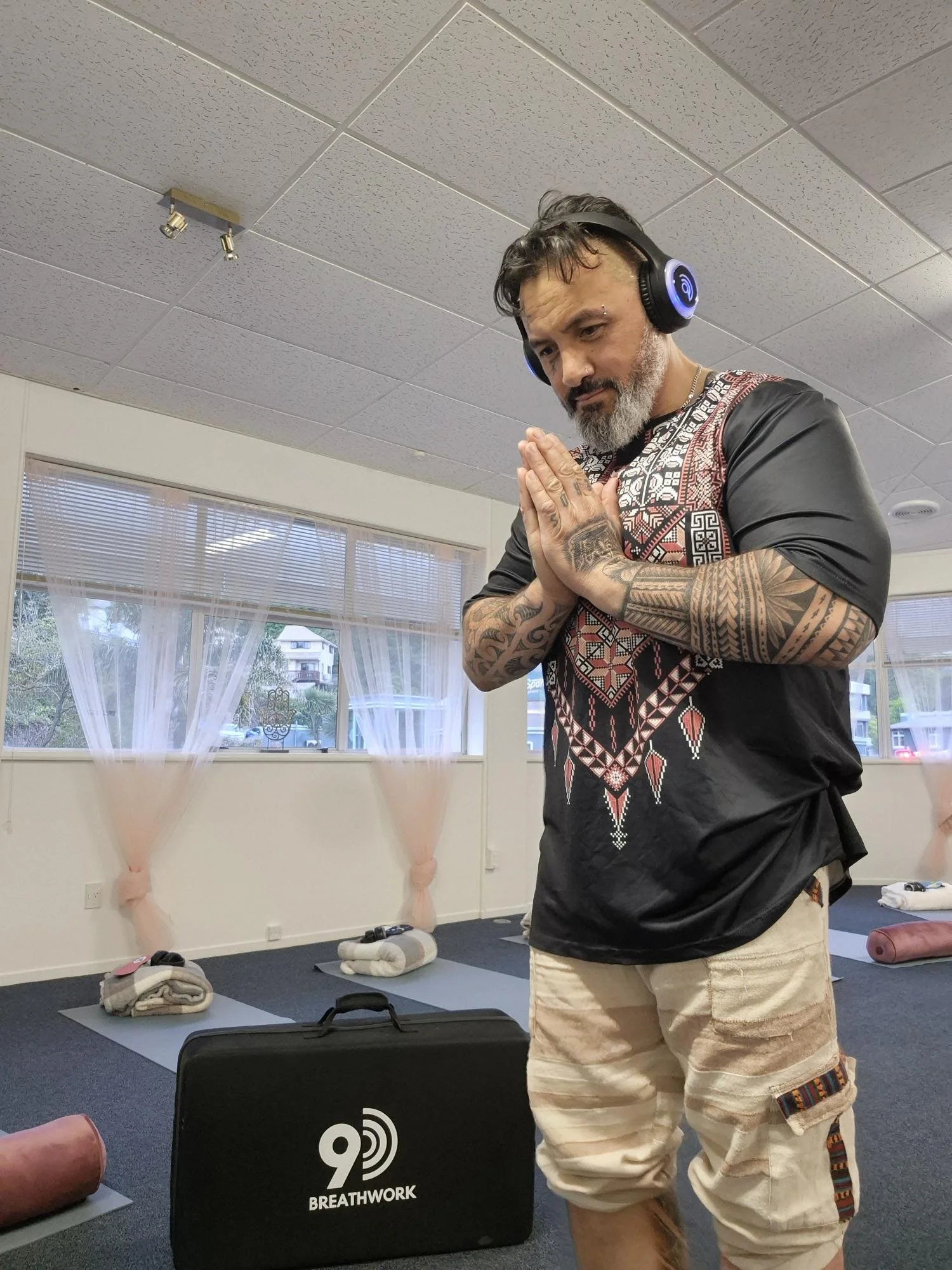 Man with tattoos and beard practicing yoga with hands in prayer position in a yoga studio, wearing headphones, surrounded by yoga mats, blankets, and yoga props.