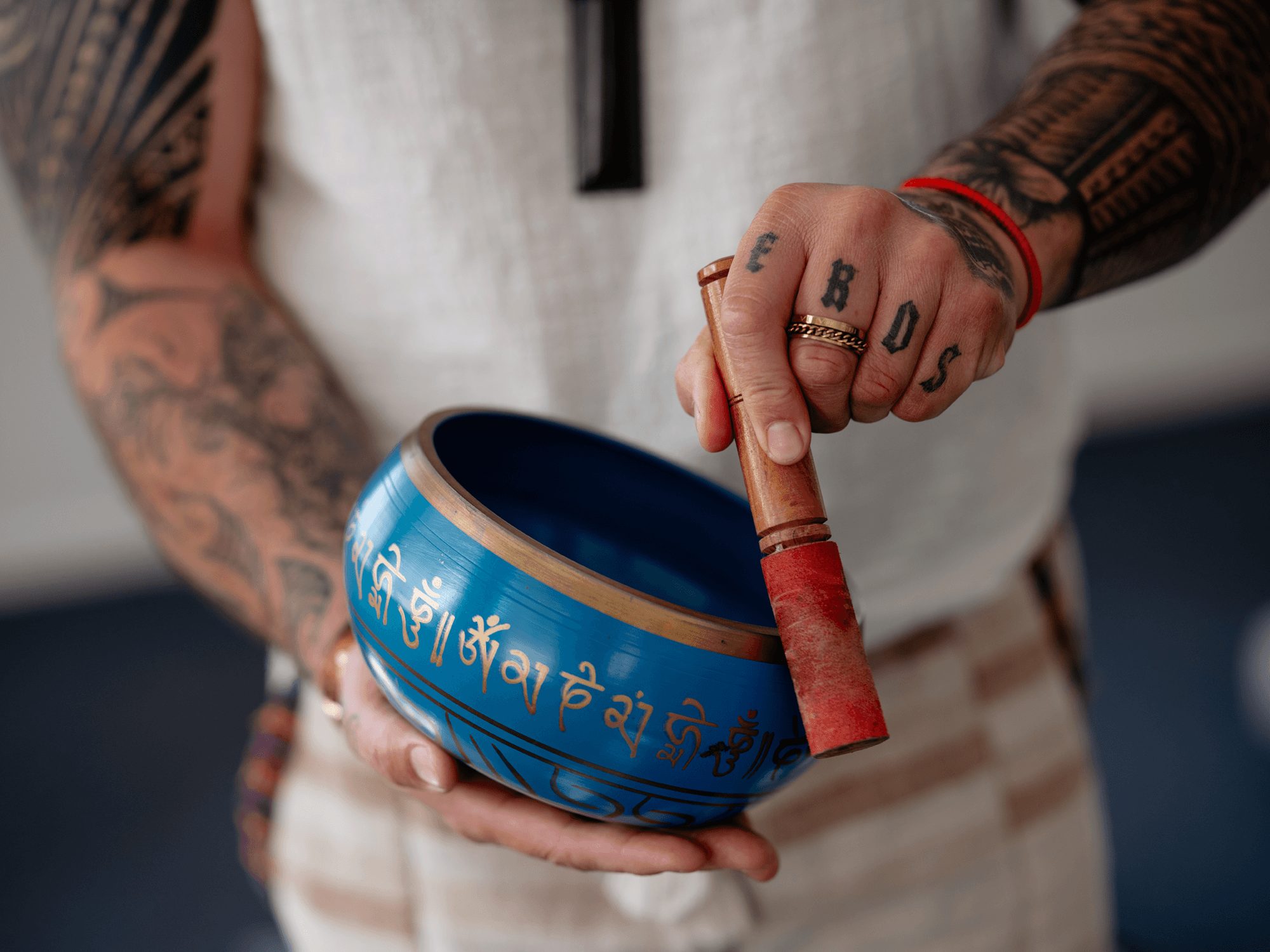 A person with tattoos on their arms and fingers holding a large blue can of Pringles potato chips, with some script writing in a foreign language on the can. The person is also gripping a red-handled wooden tool.