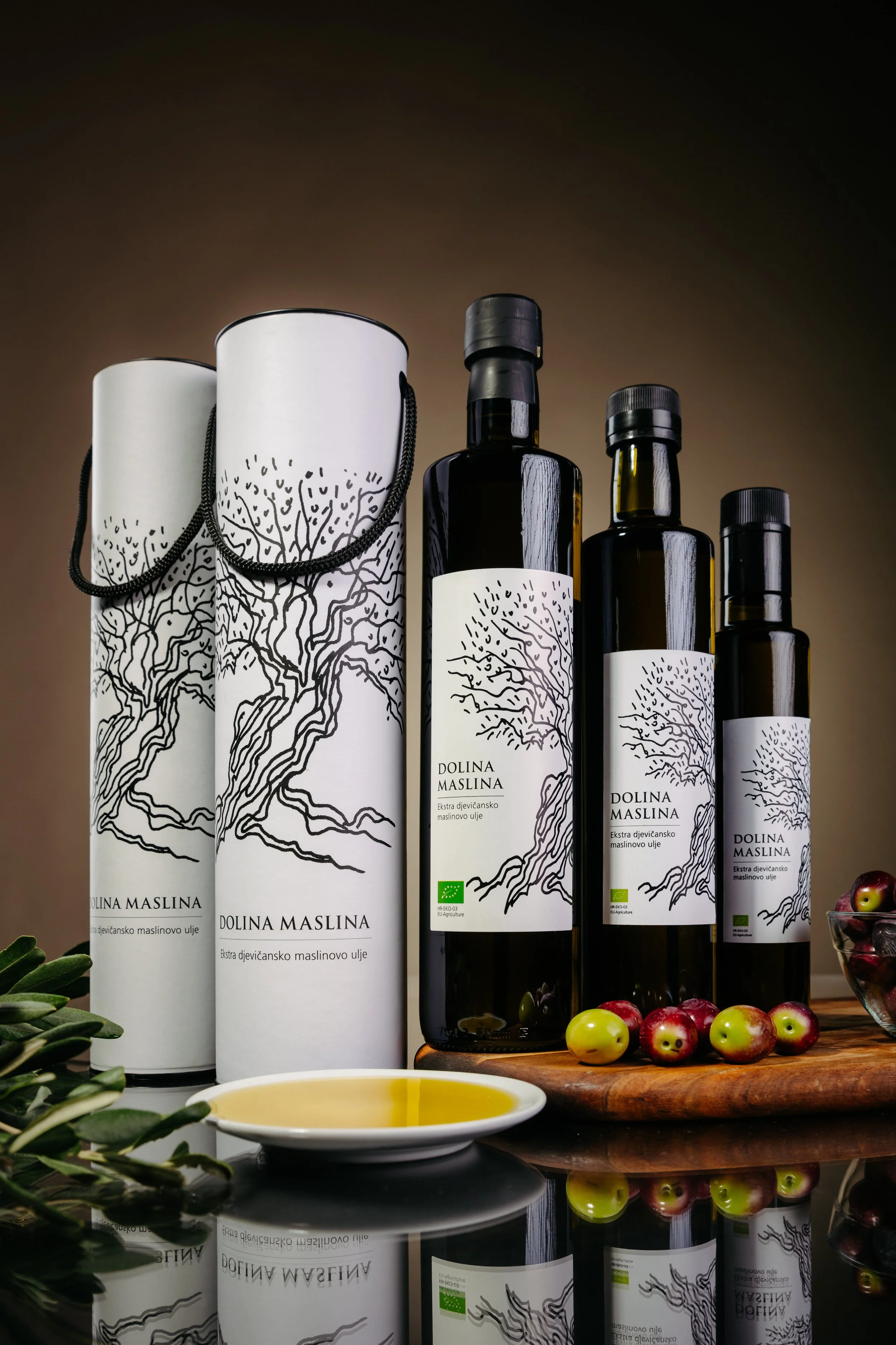 Several bottles of olive oil with design labels, some wrapped in white paper, sit on a reflective surface with a bowl of olive oil and fresh olives nearby, and leafy greens in the foreground.