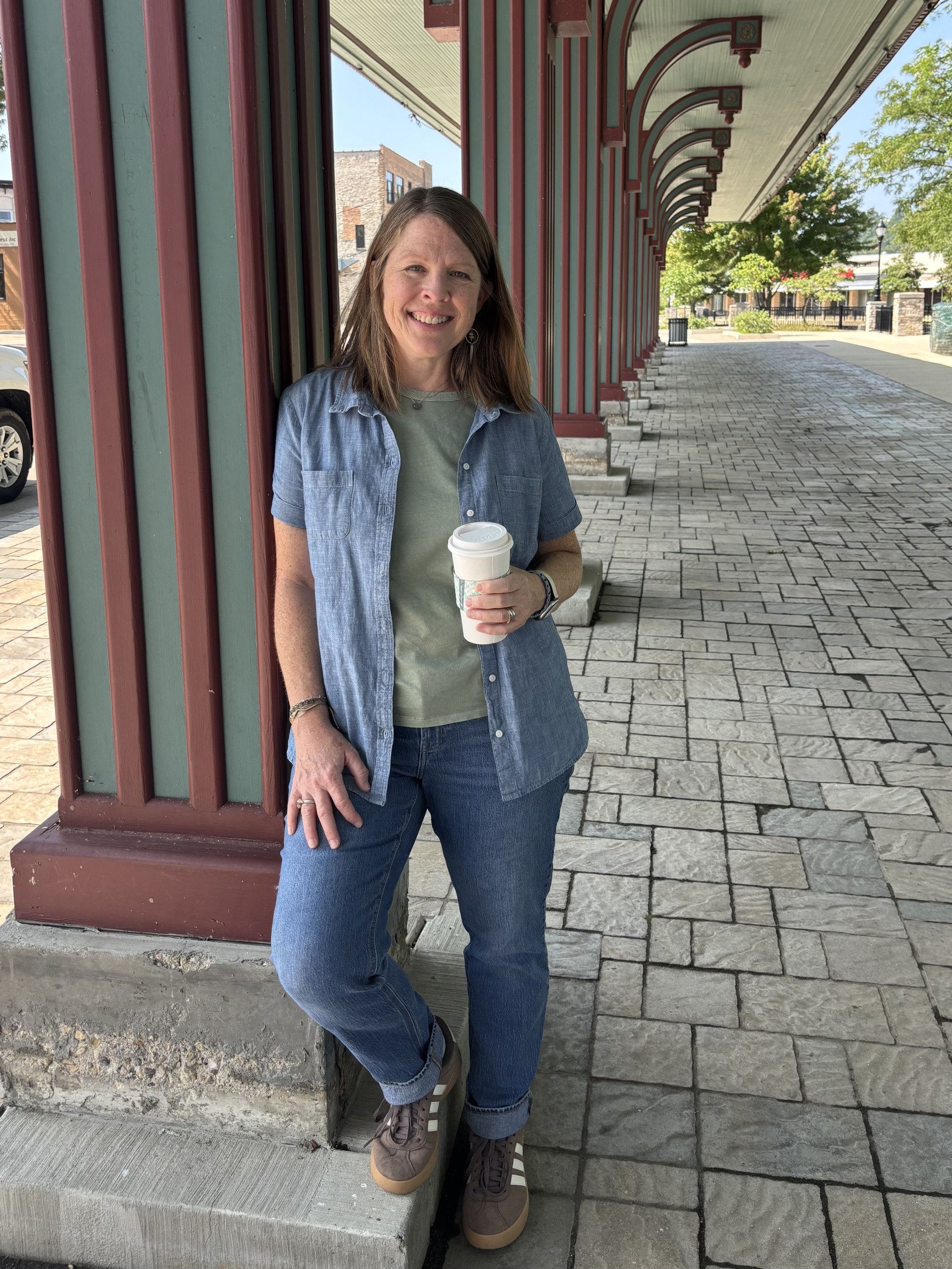 A woman standing next to an exterior wall of a building, holding a coffee cup, smiling, wearing a denim shirt, gray shirt, blue jeans, and sneakers, outside on a sunny day.