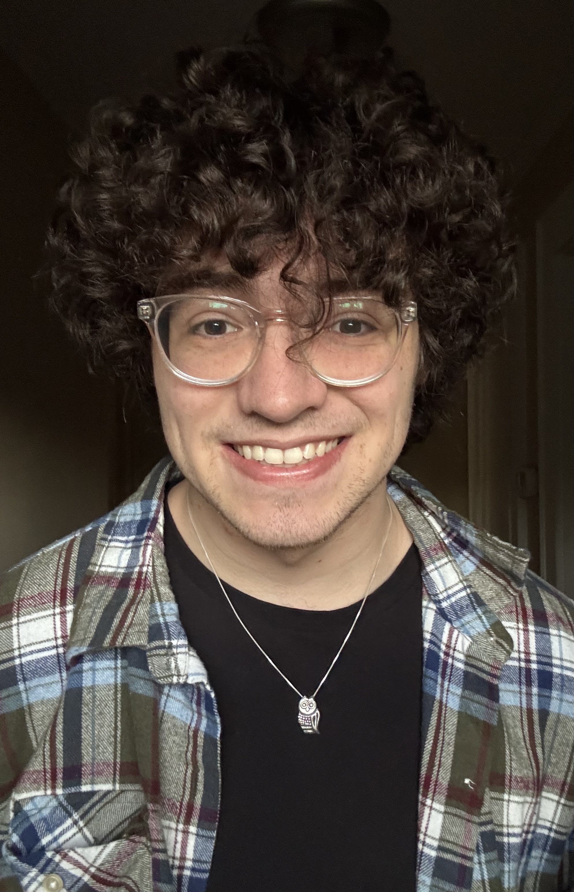Close-up of a smiling person with curly hair, wearing glasses, a plaid shirt, and a owl-shaped necklace.