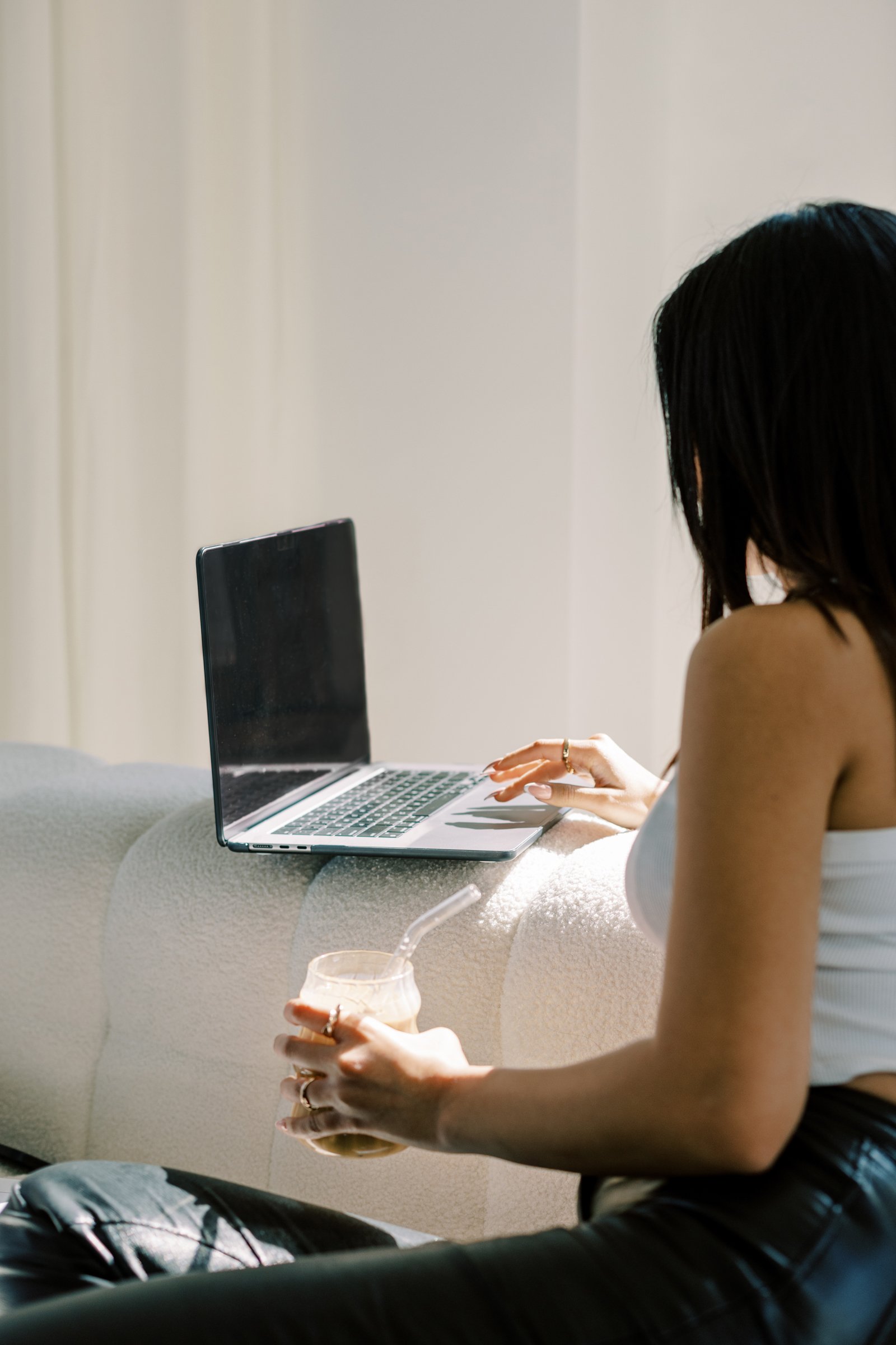 A woman with dark hair sitting on a sofa, using a laptop with a black screen, holding a cold beverage in a clear plastic cup with a straw.
