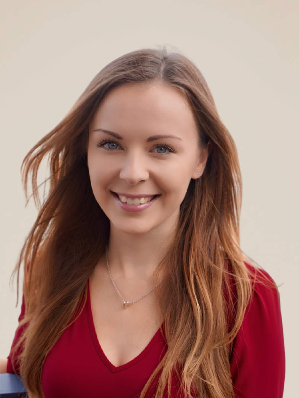 Portrait of a young woman with long, wavy auburn hair wearing a red top and a delicate necklace, smiling against a plain light background.