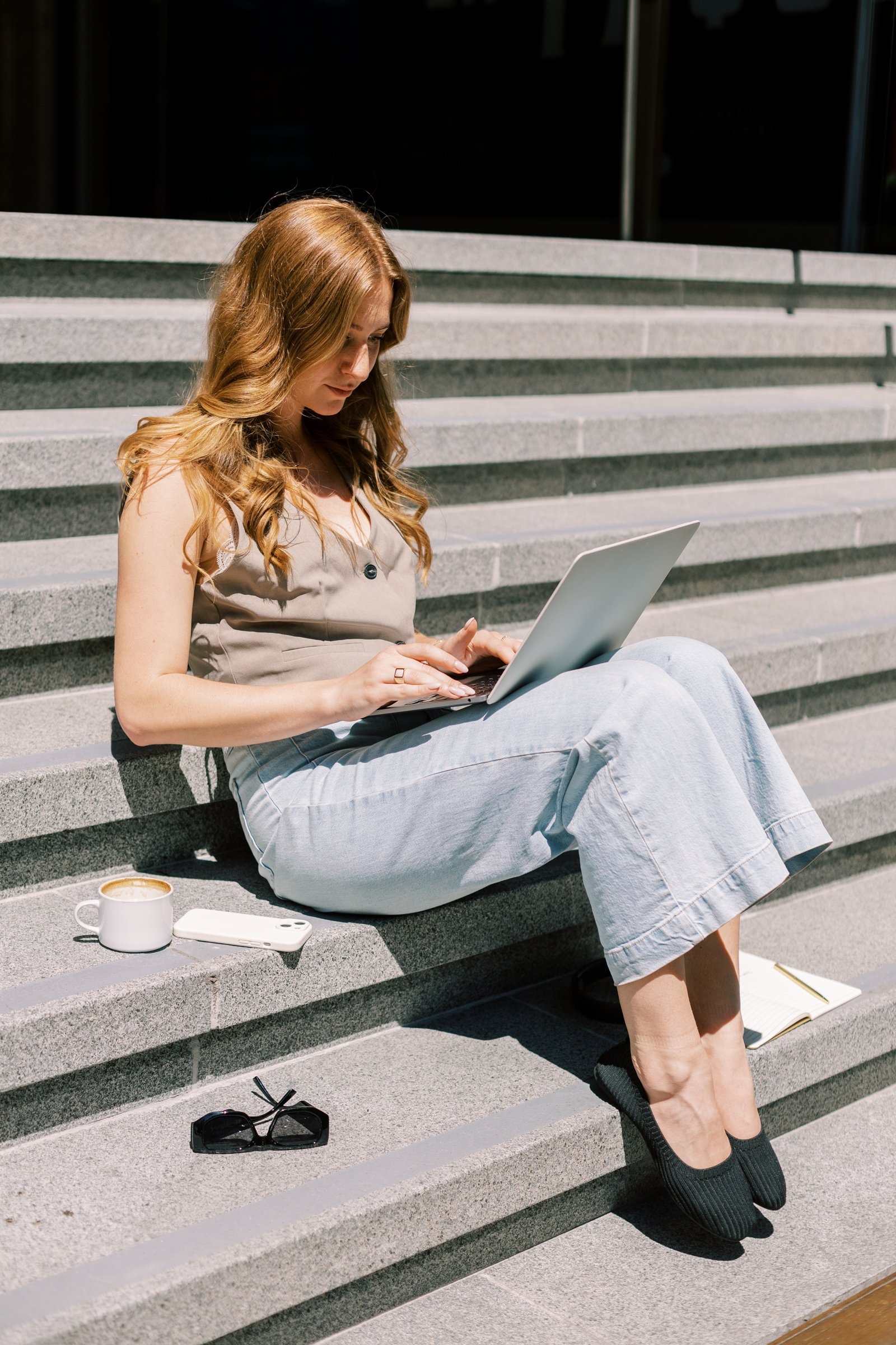 A young woman with long wavy red hair sitting on outdoor concrete stairs, looking at a laptop. She is wearing a beige sleeveless top, light blue jeans, and black slip-on shoes. A coffee mug, a white smartphone, a pair of sunglasses, a notebook, and a pen are placed on the stairs beside her.