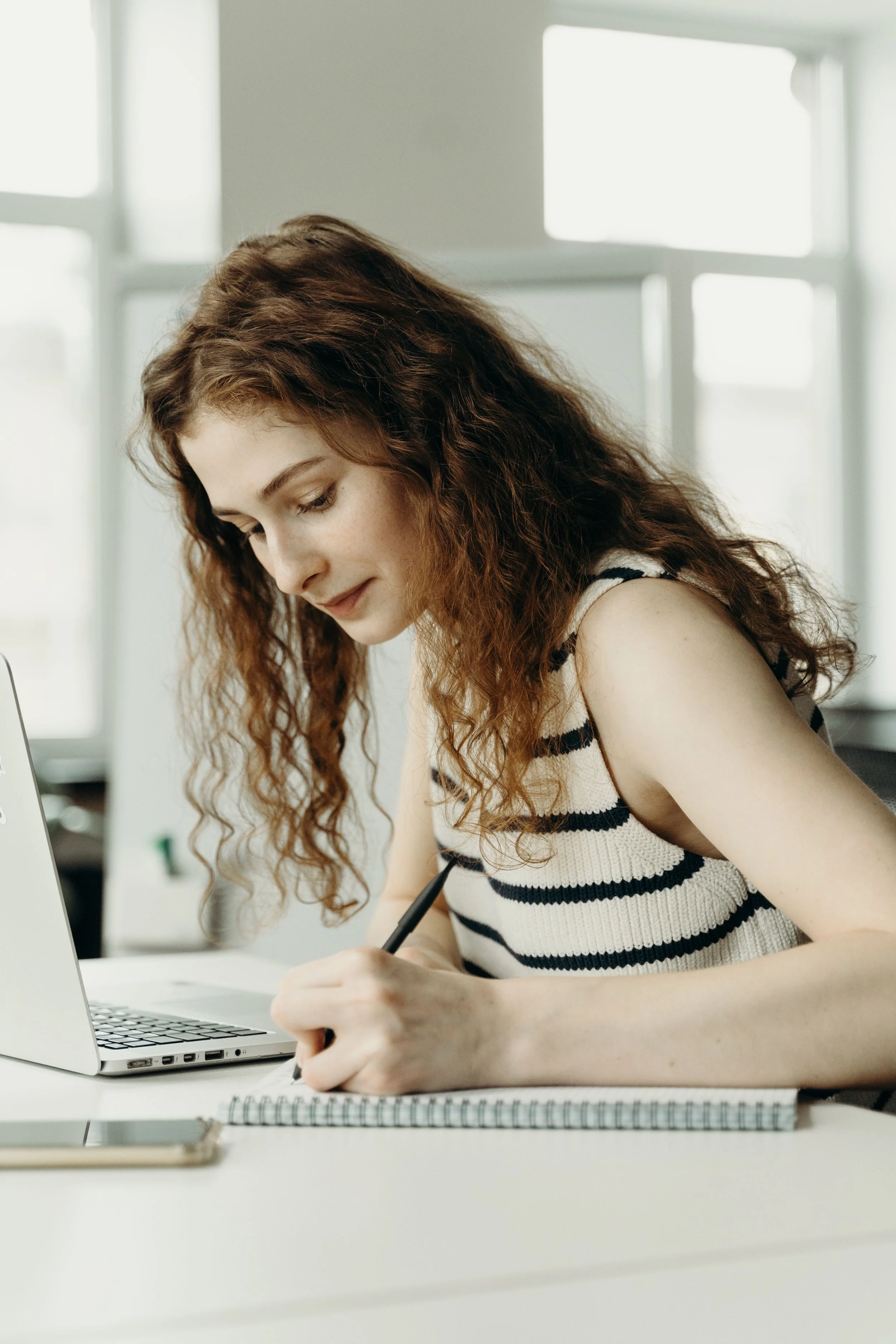 A young woman with curly red hair and light skin writing in a notebook at a desk with a laptop nearby, in front of a window with white frames.