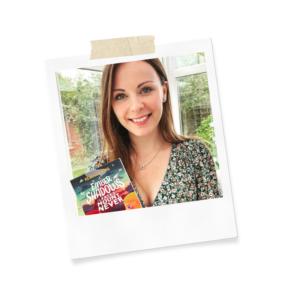A smiling woman with shoulder-length brown hair holding a book titled 'Ember Shadows' in a cozy room with large windows and green outdoor scenery in the background.