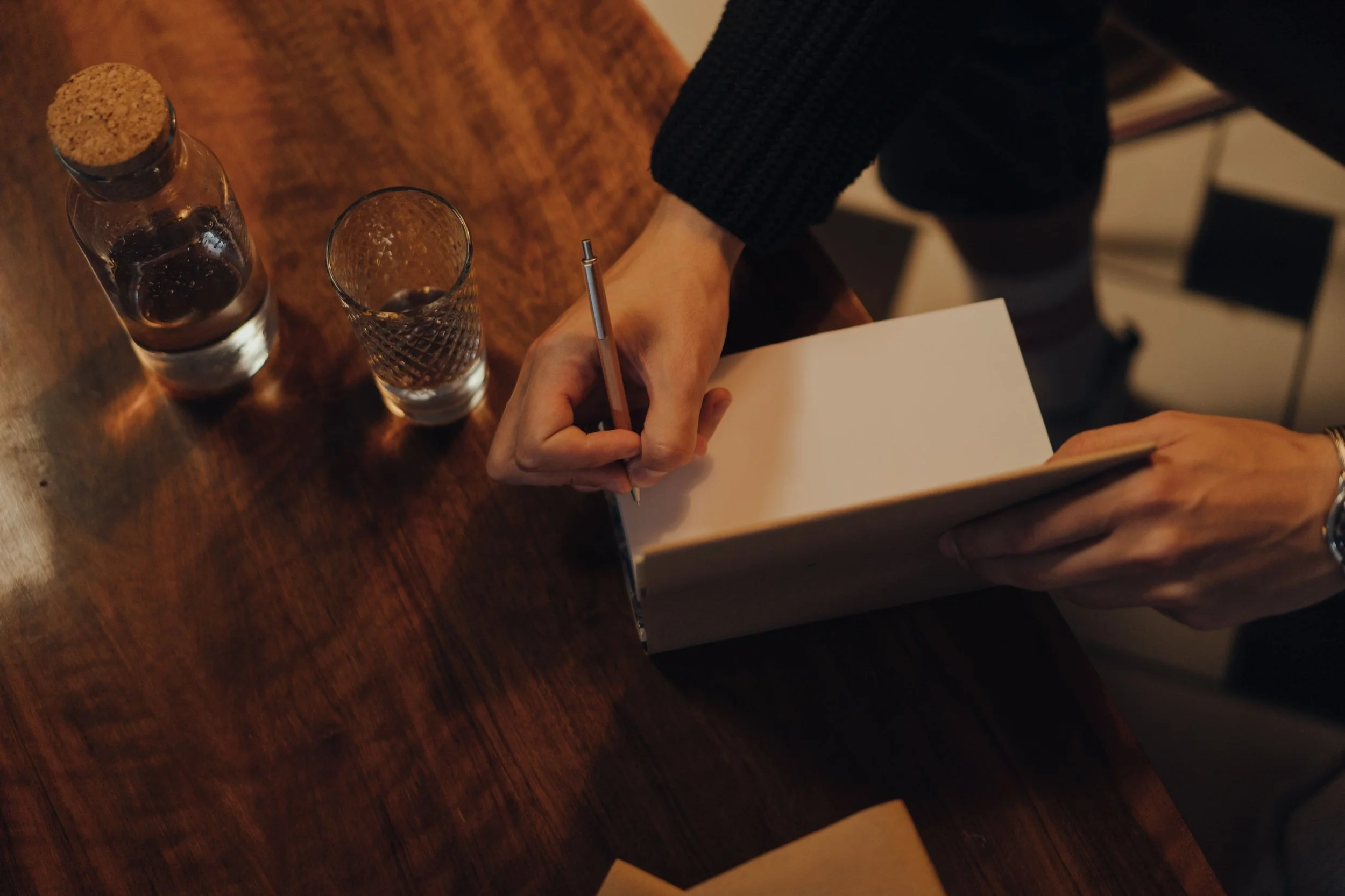 Person writing in a notebook at a wooden table with two glasses of water and a bottle of soda.