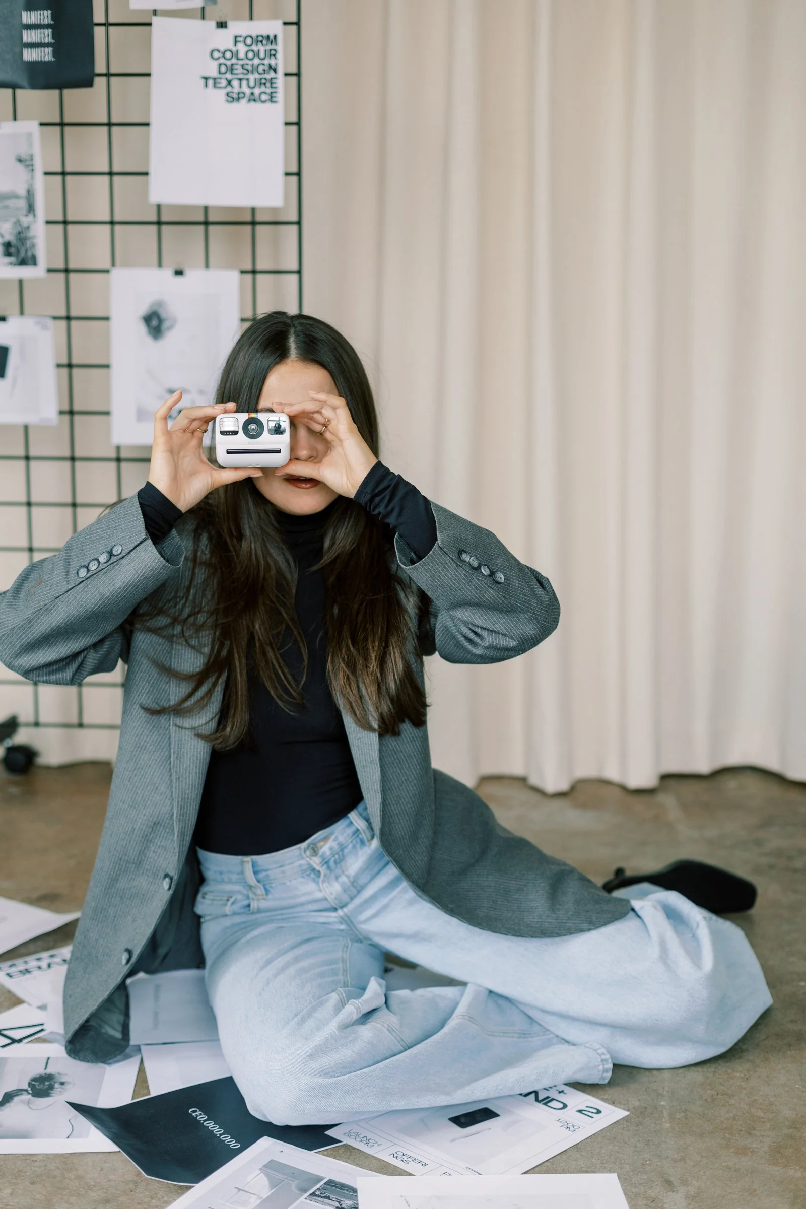 A young woman with long dark hair is sitting on the floor, wearing a gray blazer, black top, and light gray sweatpants. She is holding a small camera or device up to her face, using it as a prop to look through. There are various printed papers, magazines, and posters scattered around her on the floor, with some documents on a table nearby. The backdrop features a cream-colored curtain and a grid display with papers and images pinned to it.