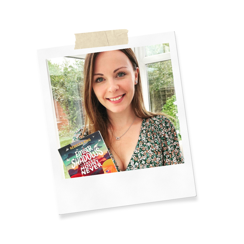 A young woman with brown hair and blue eyes smiling in a room with a window showing a garden outside, holding a book titled "Ember Shadow" by Rebecca King.