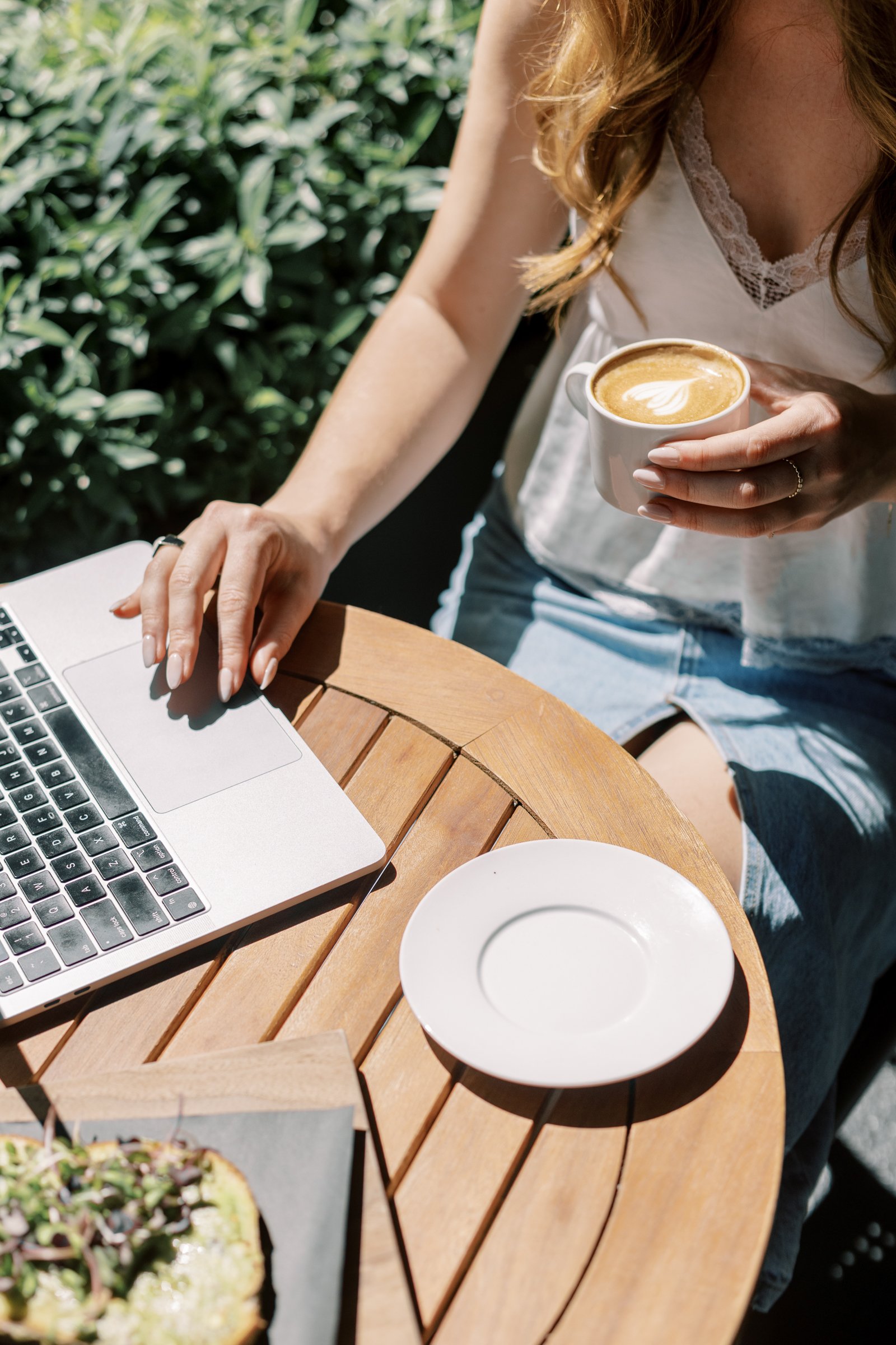 A person is sitting at a wooden outdoor table, working on a laptop, holding a cup of coffee with latte art, wearing a white top, with a plate and food in the foreground.