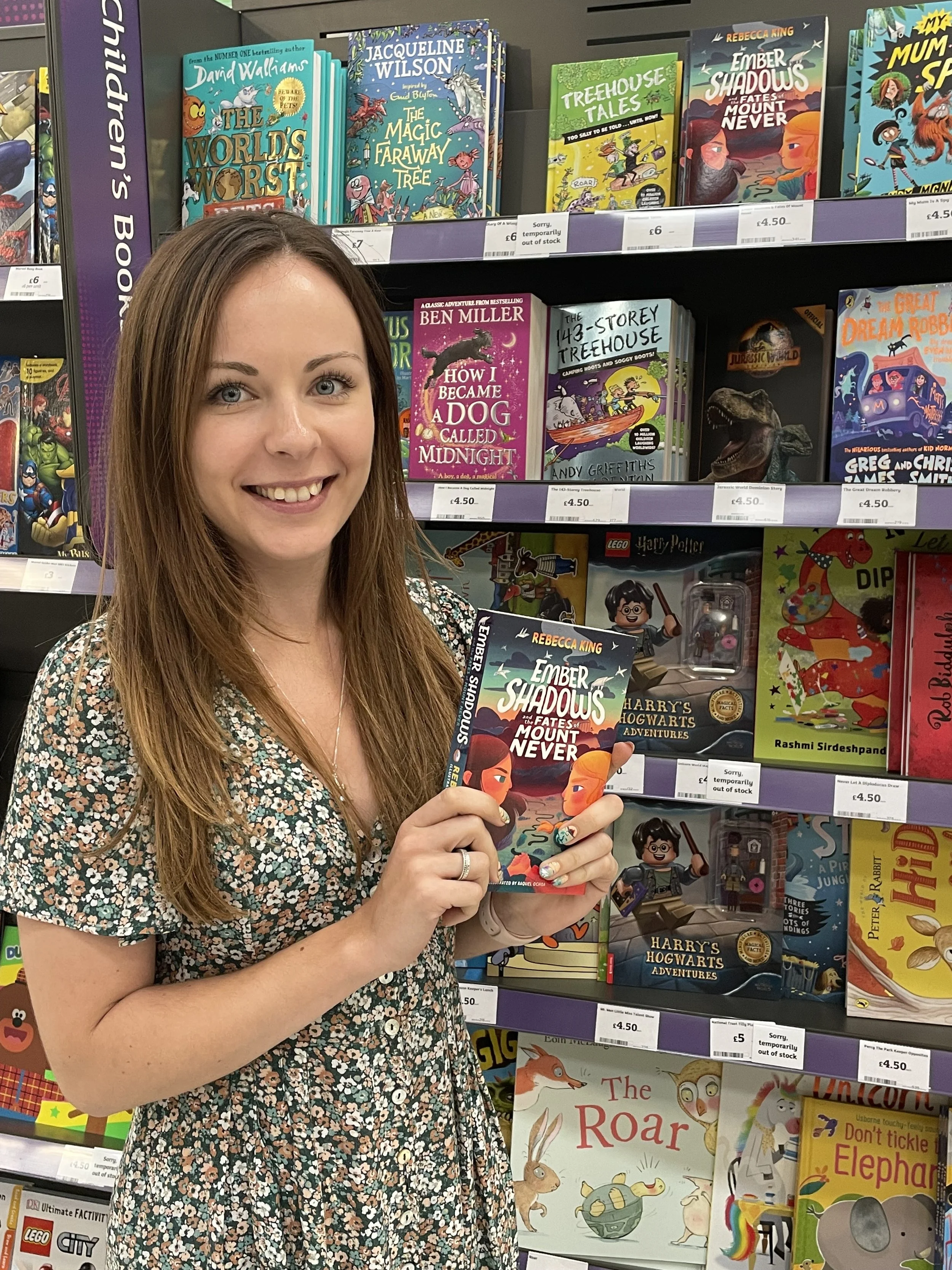 A woman with long red hair smiling and holding a children's book titled 'Ember Shadows and the Fates of Mount Never' in a bookstore. Bookshelves behind her display various children's books, including titles about animals, magic, and adventures.