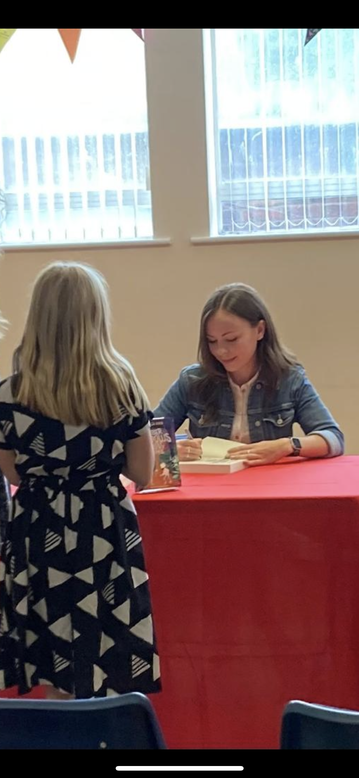 A young woman with shoulder-length brown hair, wearing a denim jacket and a watch, signs a book at a table covered with a red cloth. A girl with long blonde hair, wearing a black and white geometric patterned dress, stands in front of her, holding a book.