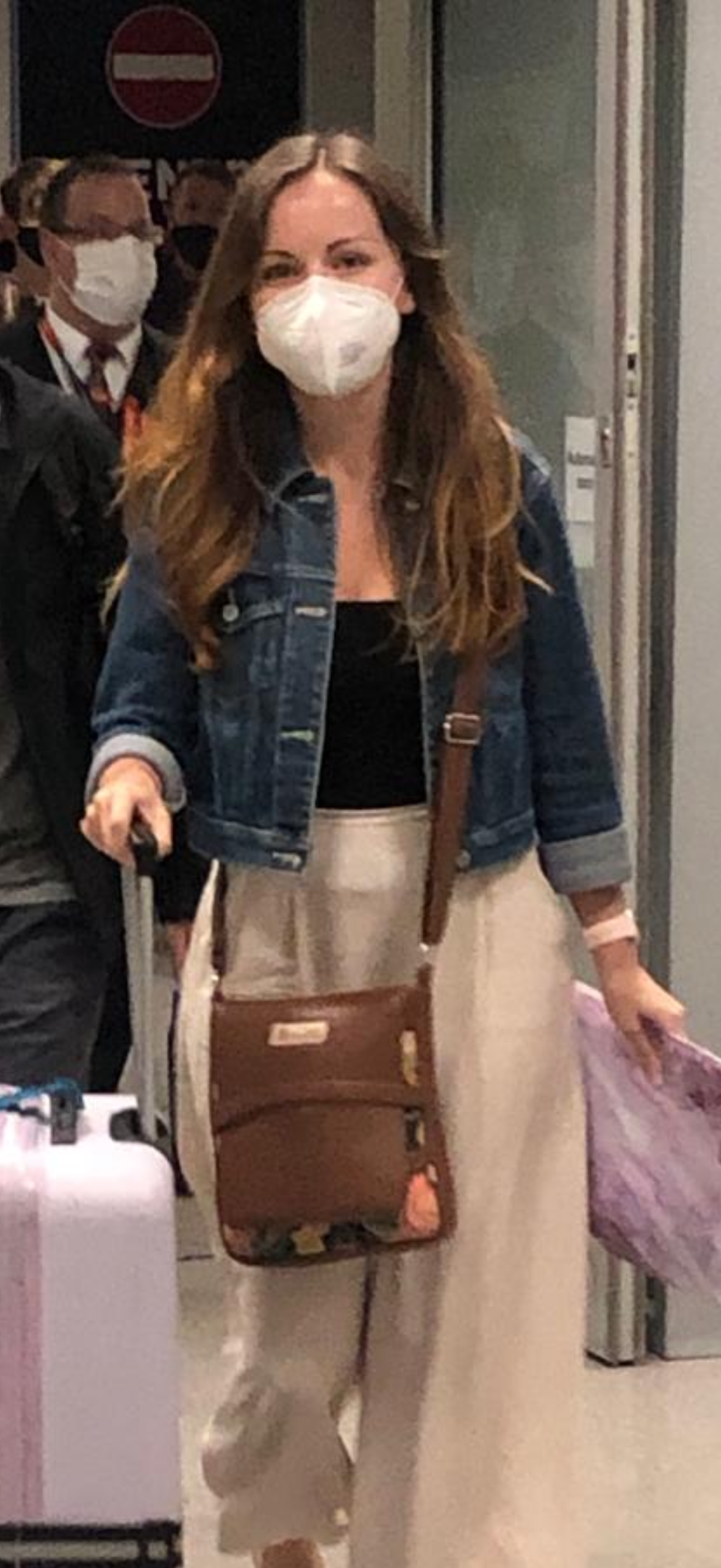 A woman wearing a face mask, denim jacket, black top, and beige pants holding a small suitcase, walking through an airport.