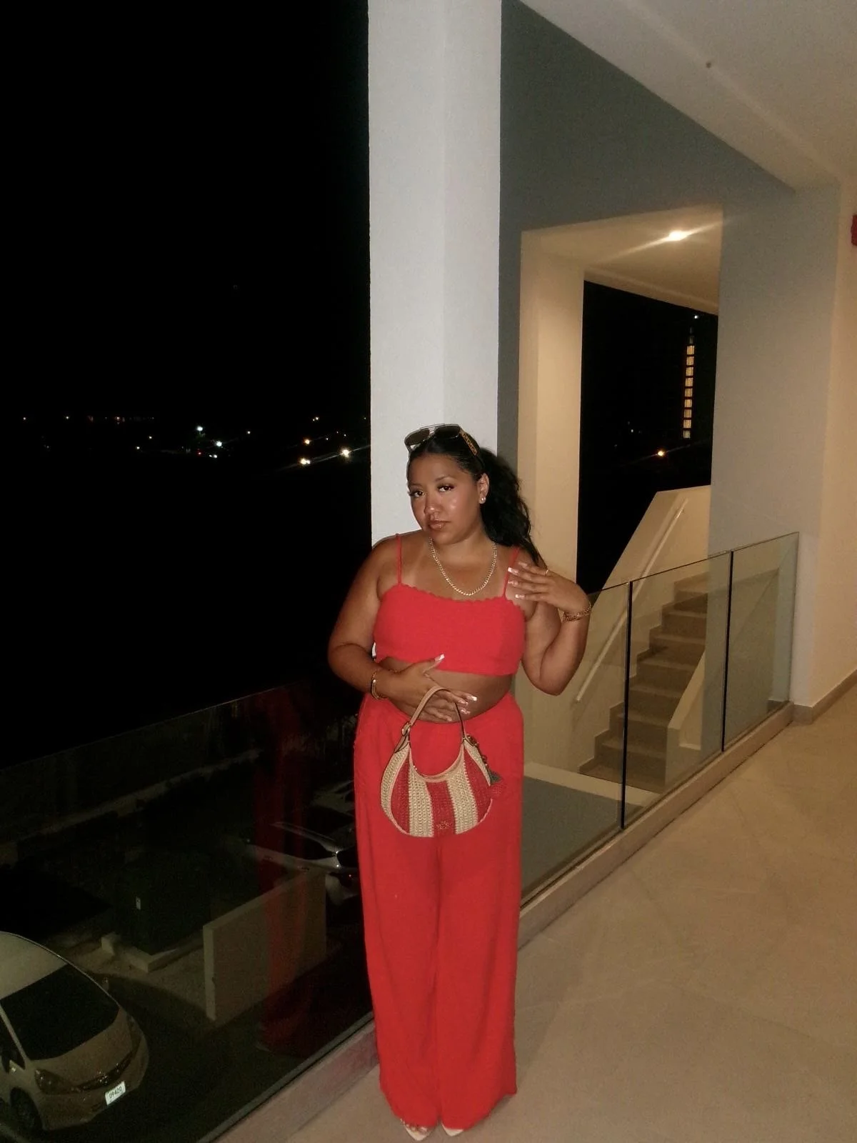 A woman standing indoors near a balcony at night, wearing a red two-piece outfit, holding a beige and red striped handbag, with sunglasses on her head and a necklace.