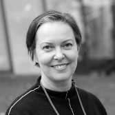 Black-and-white portrait of a woman smiling, with short hair and a necklace, outdoors in a park or garden setting.