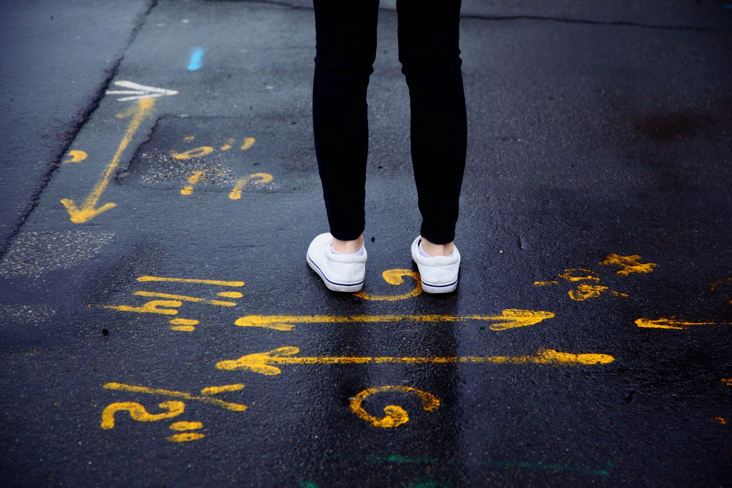 Person standing on asphalt street with yellow arrows in many directions on the ground.