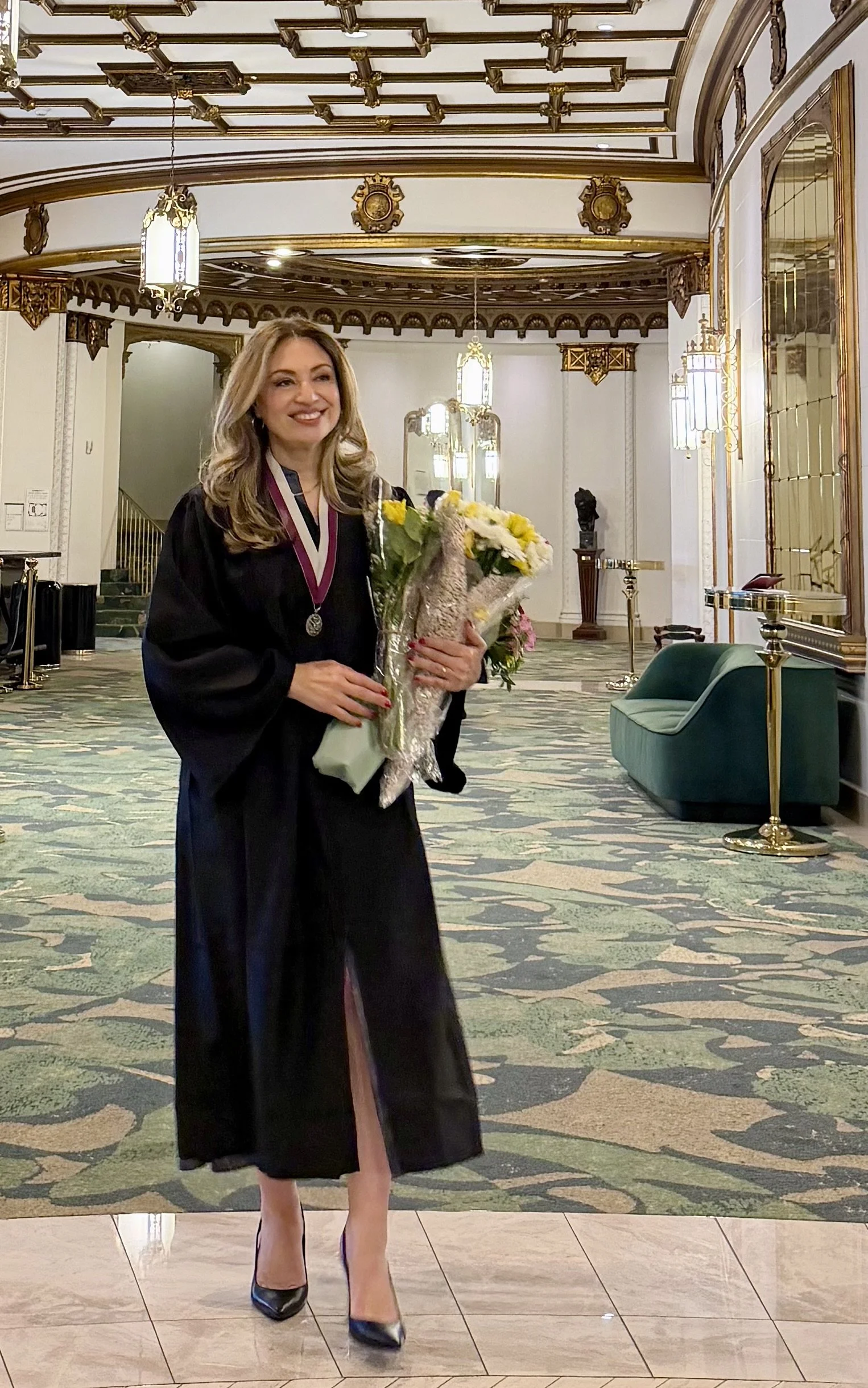 A woman in a black academic gown holding a bouquet of flowers and wearing a medal, standing in an ornate, elegant lobby with decorative gold accents, chandeliers, and a patterned carpet.