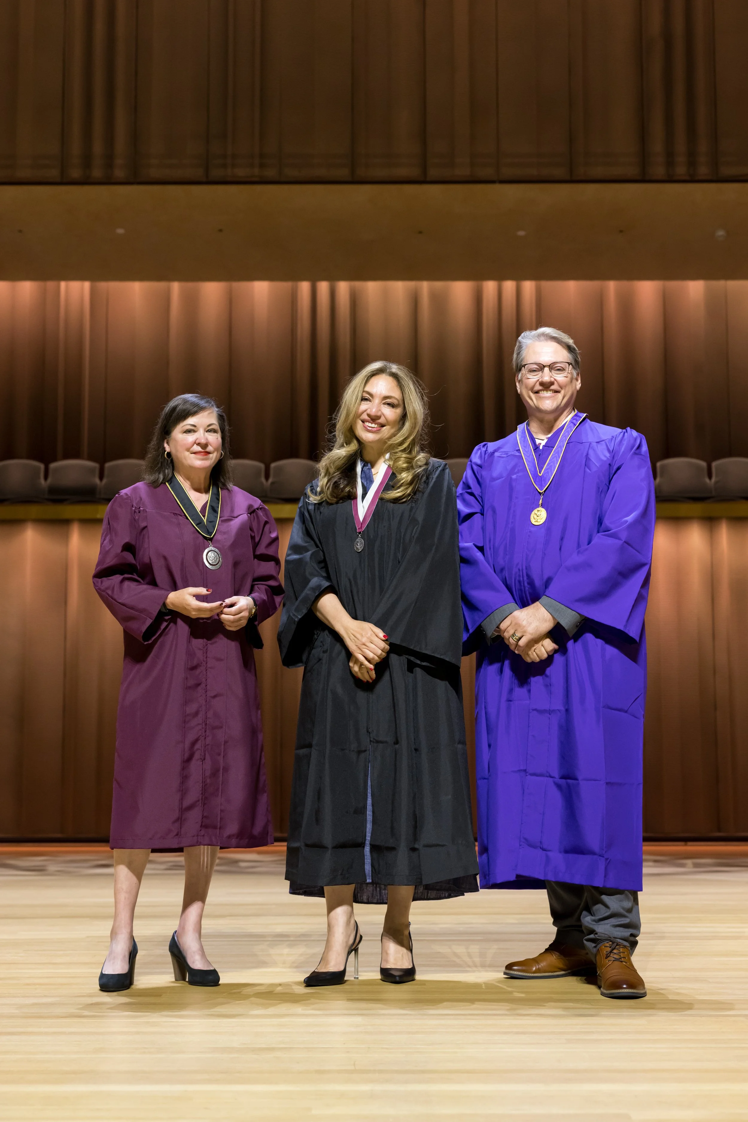 Three individuals wearing academic regalia standing on a stage, smiling, in an auditorium with wooden paneling and empty chairs in the background.