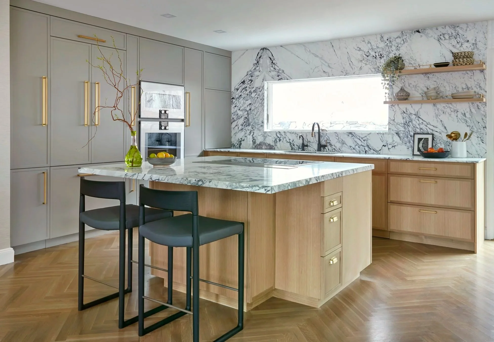 Modern kitchen with a marble island, light gray cabinetry with gold handles, a large marble backsplash, and two black chairs at the island. There are open shelves with dishes and a window above the sink.