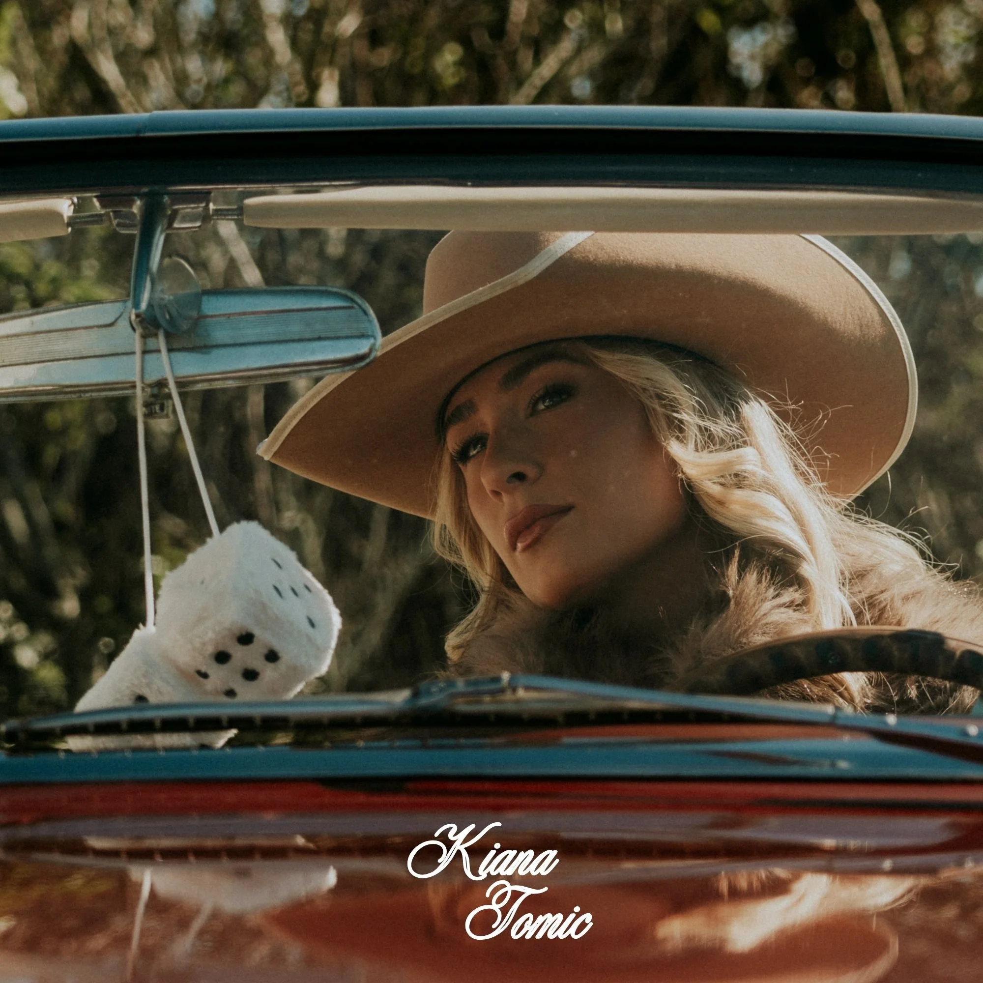 Close-up of a woman's face with blonde hair and a wide-brimmed hat, looking through a vehicle's sunroof with trees in the background. A straw hat with a ribbon is also visible. The image includes overlay text that says "Kiana & Tomic."
