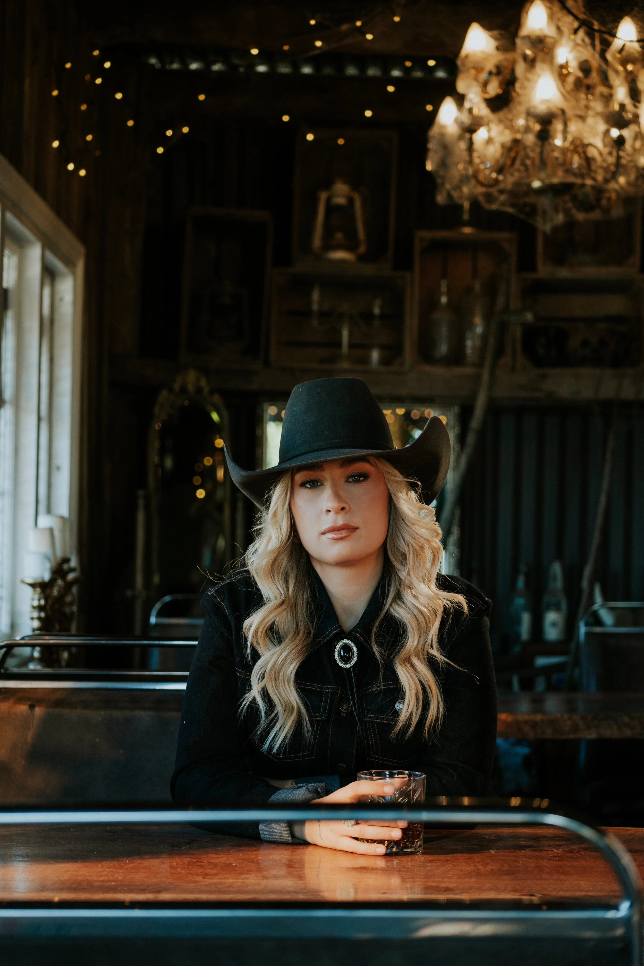 A woman with long blonde hair wearing a black cowboy hat and black clothing sitting at a bar counter holding a glass of dark beverage. The background features dim lighting, a chandelier, wooden shelves, and a rustic interior.
