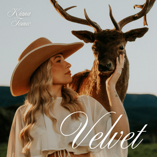 A woman with long blonde hair wearing a wide-brimmed hat and a light-colored blouse, standing beside a large reindeer with impressive antlers, in an outdoor setting with a natural landscape background.