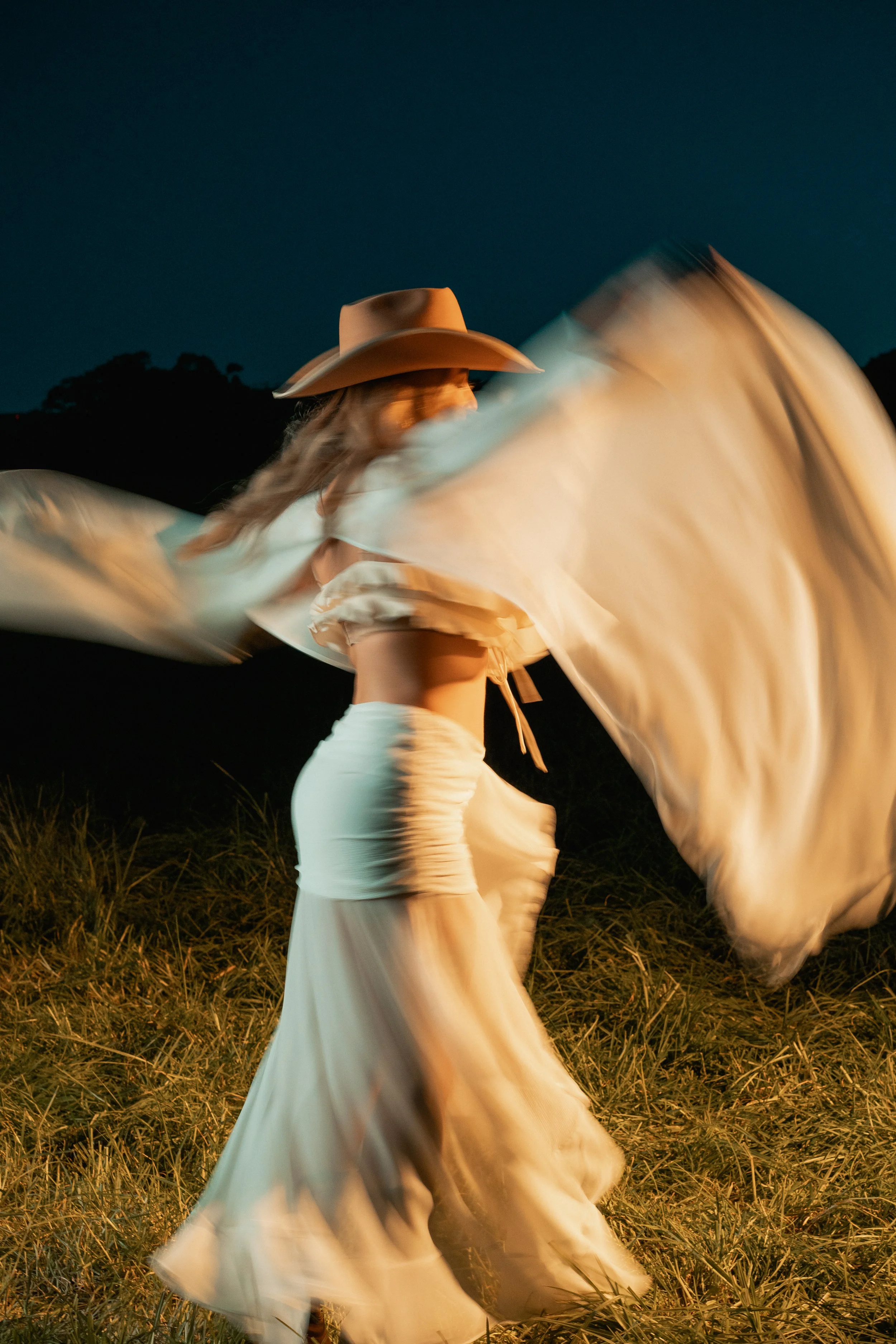 A woman wearing a cowboy hat and a white flowing dress spinning in a grassy outdoor area at night, with a dark mountain silhouette in the background.