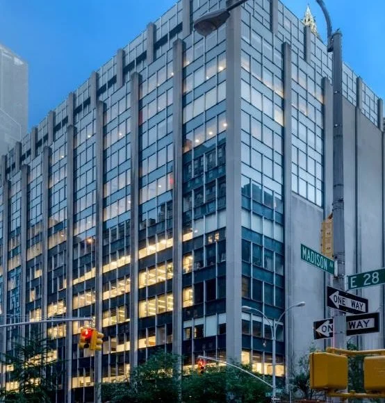 Tall modern office building with glass windows reflecting the cityscape, located at the intersection of Madison Avenue and East 28th Street, with traffic lights and street signs visible.
